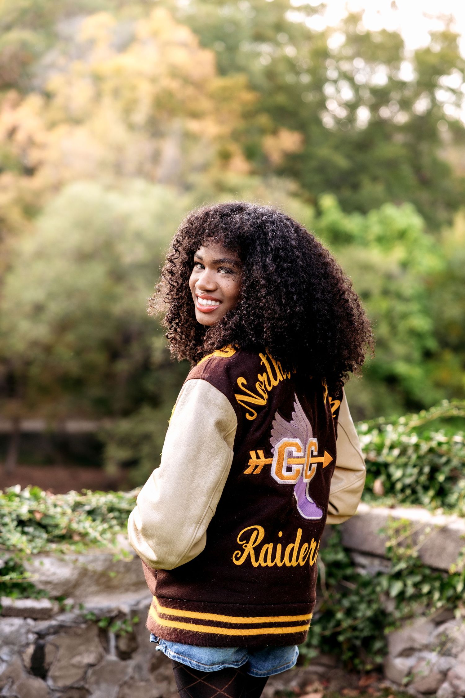 Senior girl smiles in her varsity jacket at Cranbrook House and Gardens by senior photographer Rachel Martinez in Metro Detroit