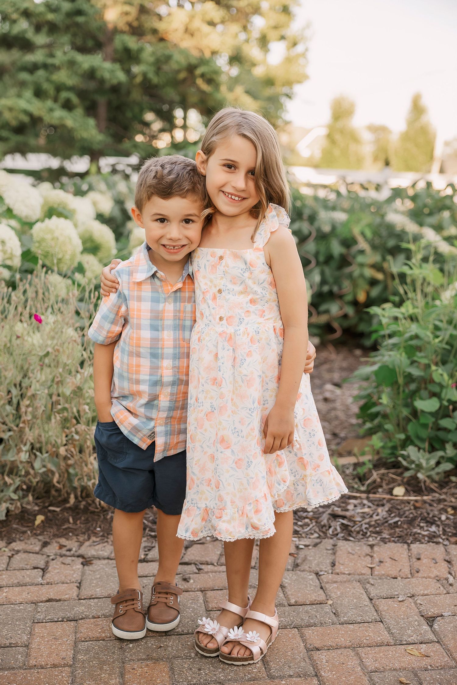 Two children pose together on brick path surrounded by garden flowers. Novi Northville Portrait Photographer