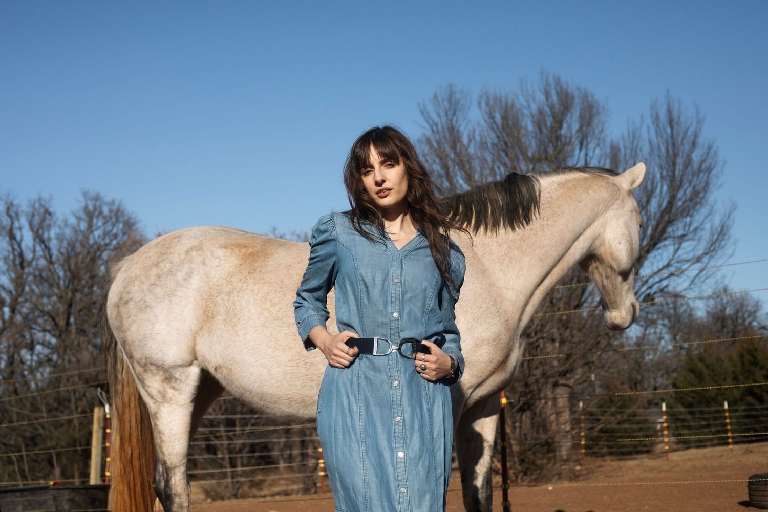 Woman in blue denim dress with belt standing beside white horse in outdoor paddock.