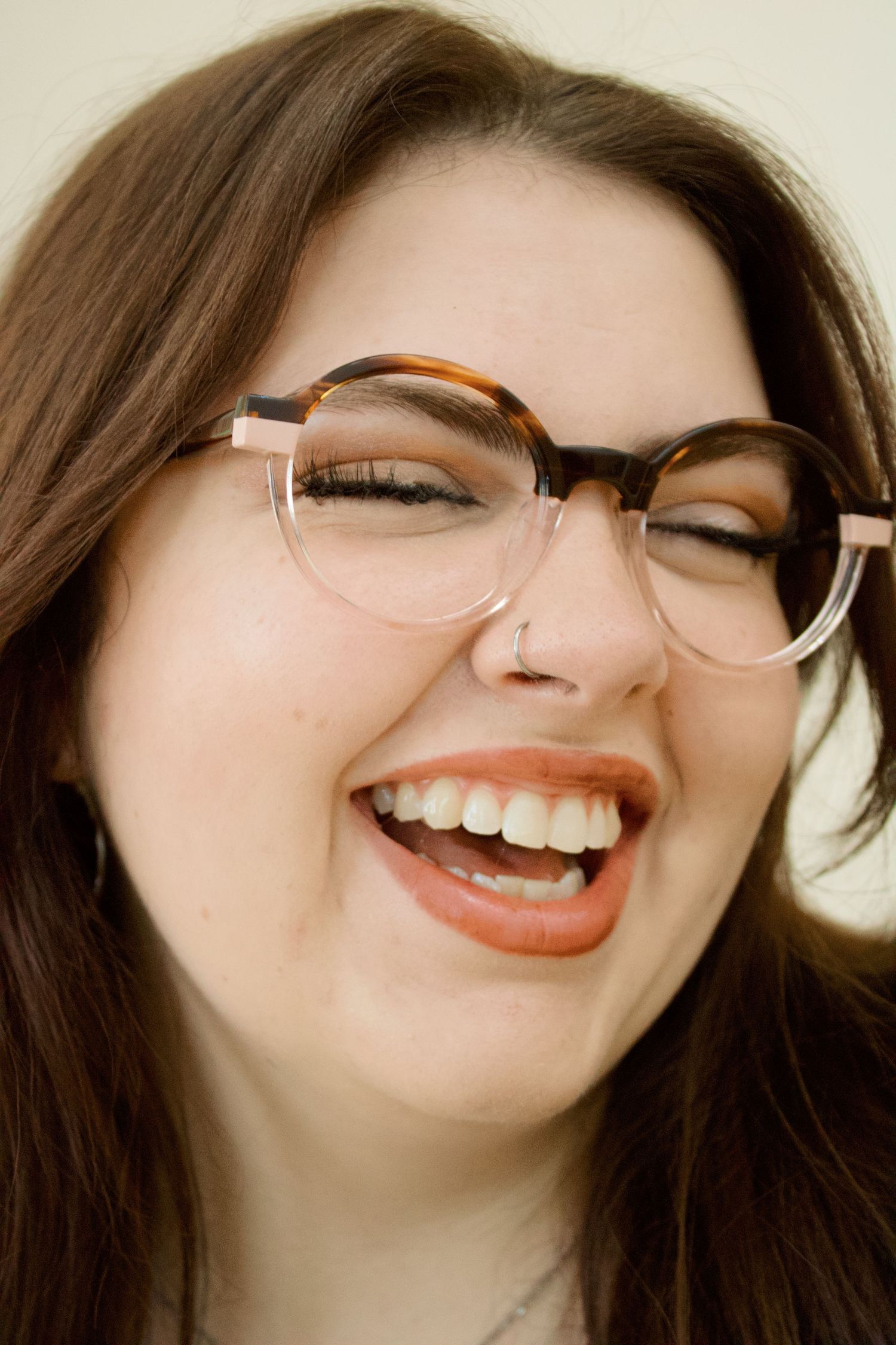 Laughing brunette woman wearing tortoiseshell glasses with pink accents against neutral background.