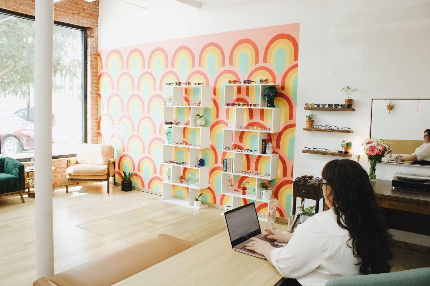 Woman working on laptop in colorful workspace with vibrant rainbow patterned accent wall and shelving.