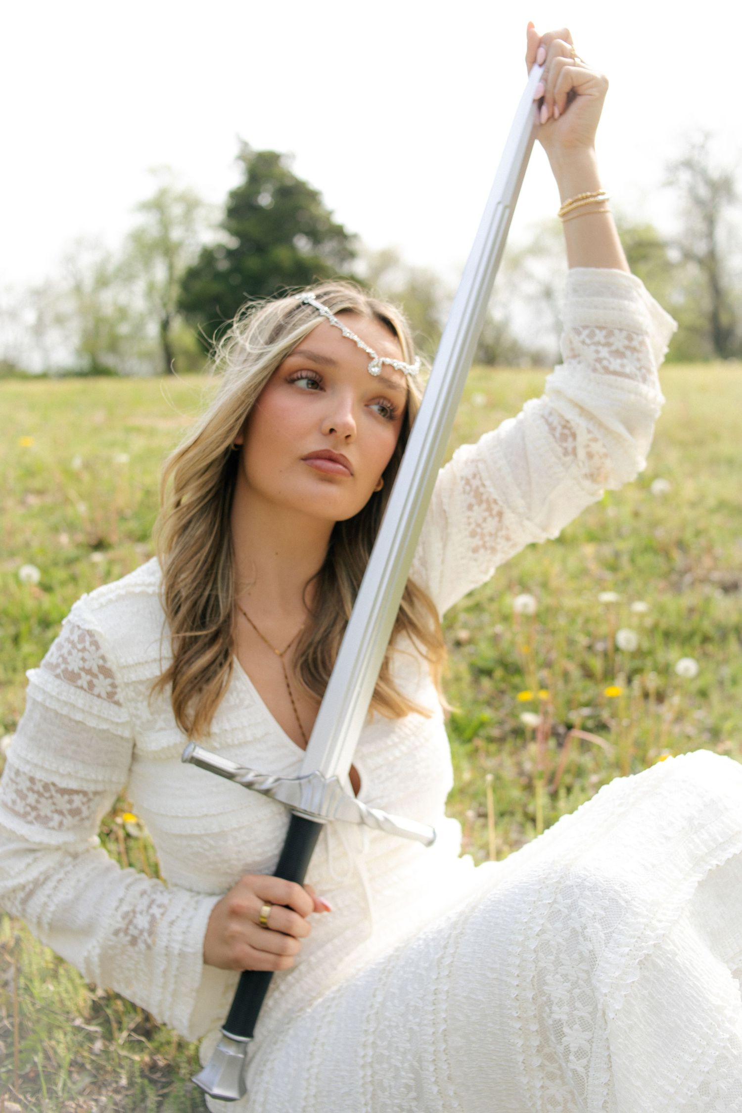 Blonde woman in white lace dress holding silver sword upright in green meadow.