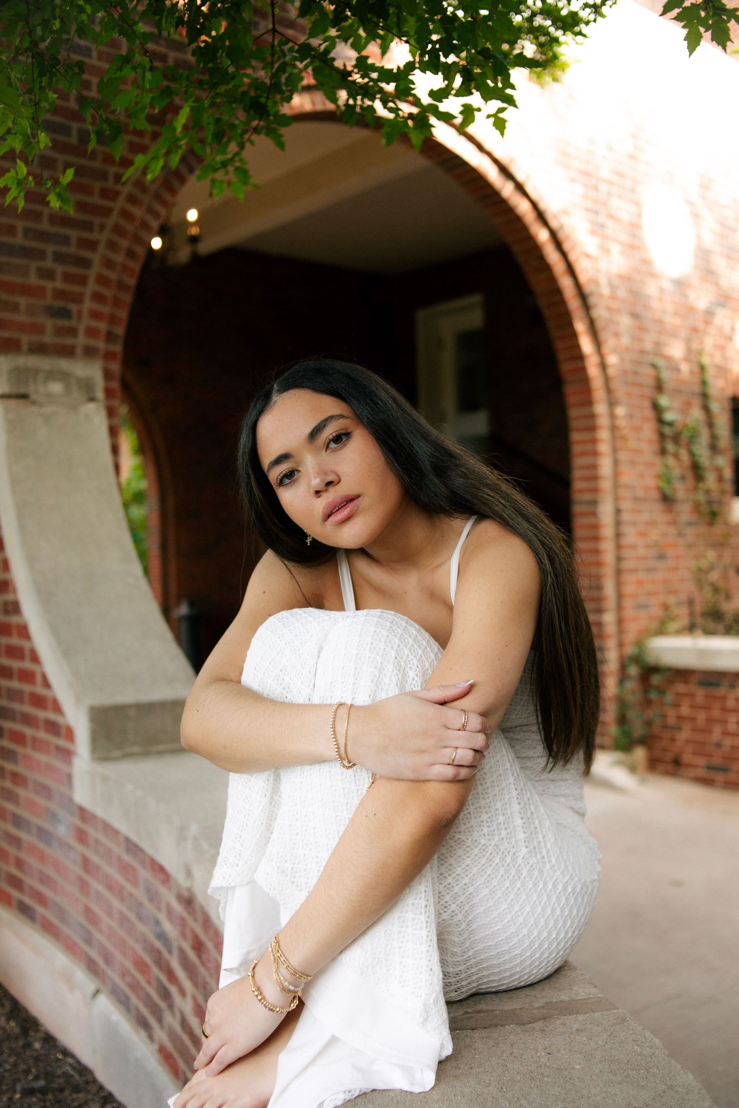 Young woman in white dress sitting under brick archway surrounded by green foliage.