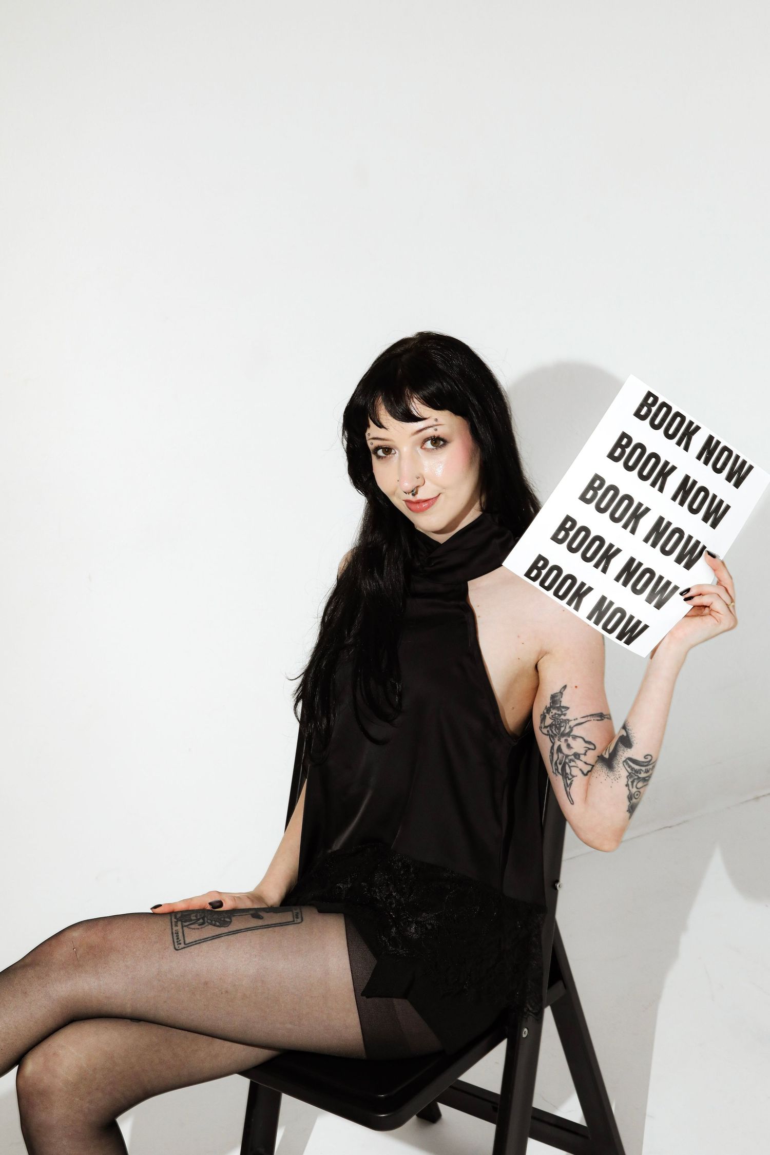 Woman in black dress sitting on chair holding a 'Book Now' sign, showcasing tattoos on arm and leg.