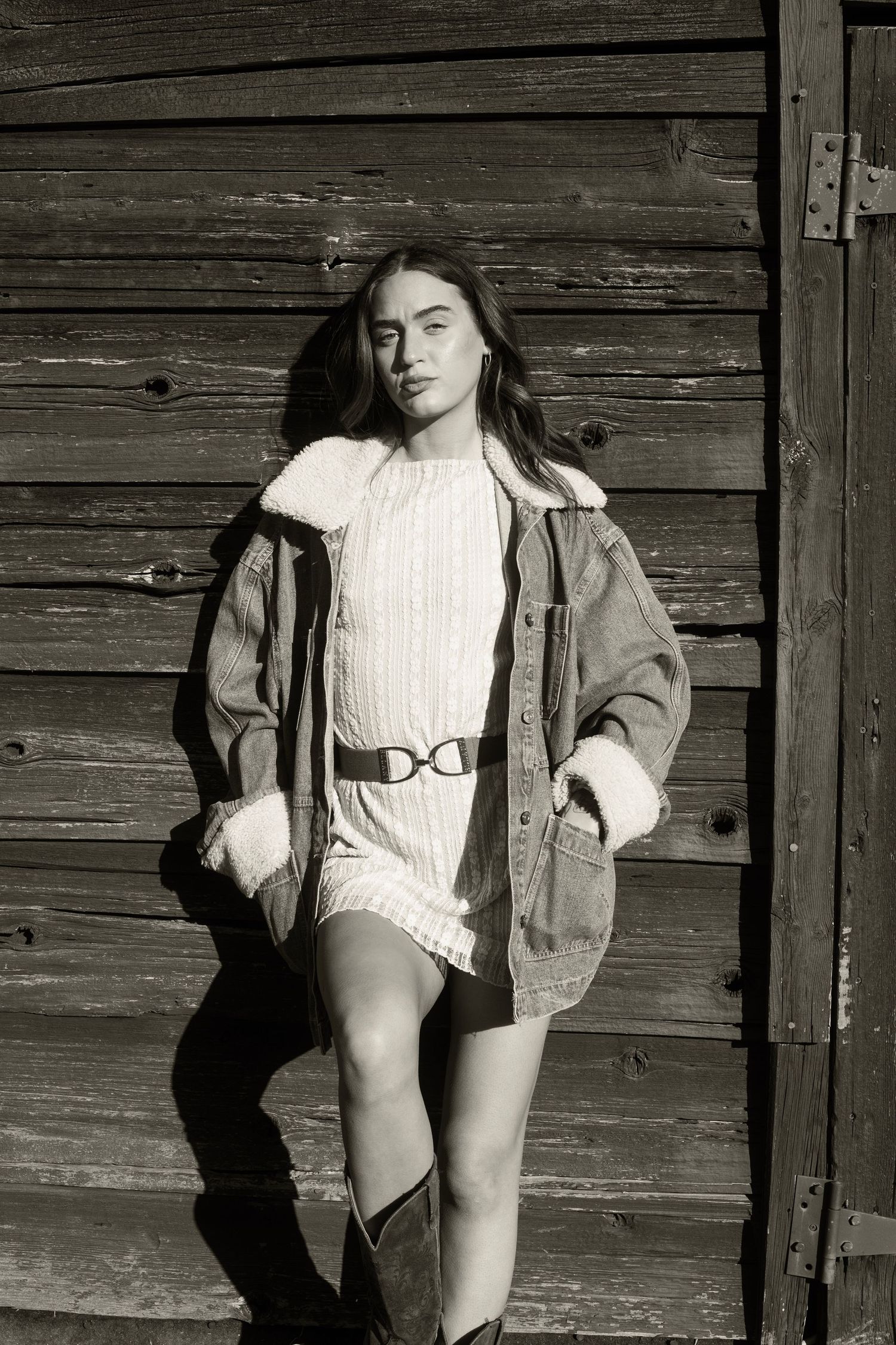 Young woman in denim jacket over white knit dress with belt and boots posed against wooden barn wall.