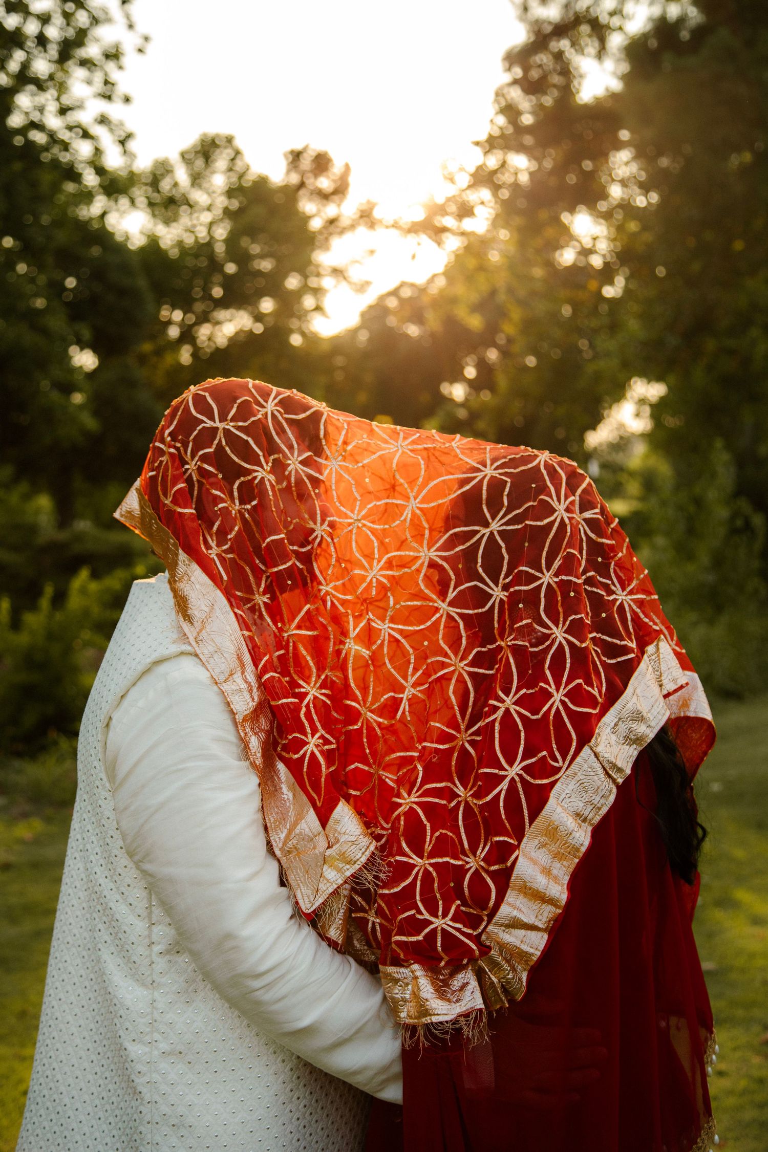Couple kissing hidden under a red embroidered dupatta veil at golden hour outdoors.
