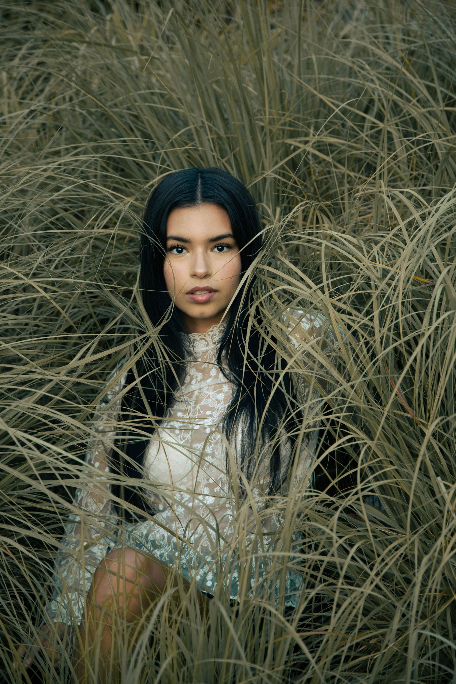 Young woman with long black hair posing among tall dry grass in a white lace top.