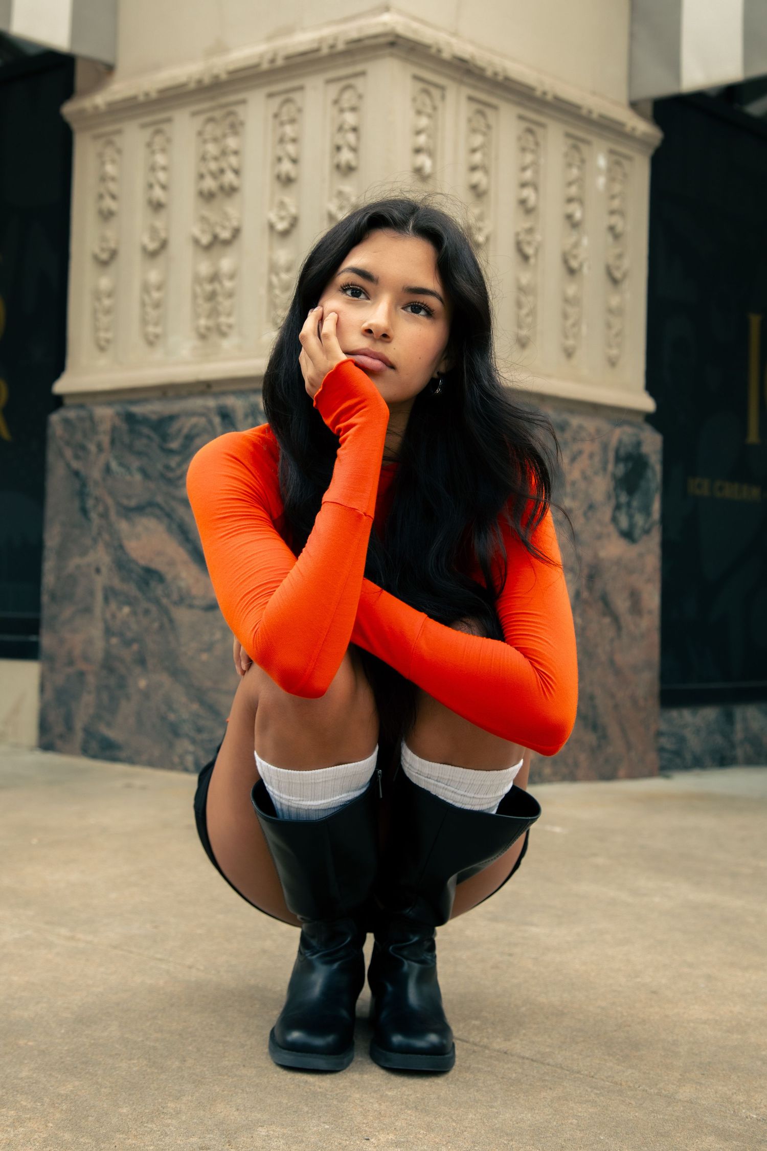Young woman in orange long-sleeve top crouching outdoors near an ornate stone building.
