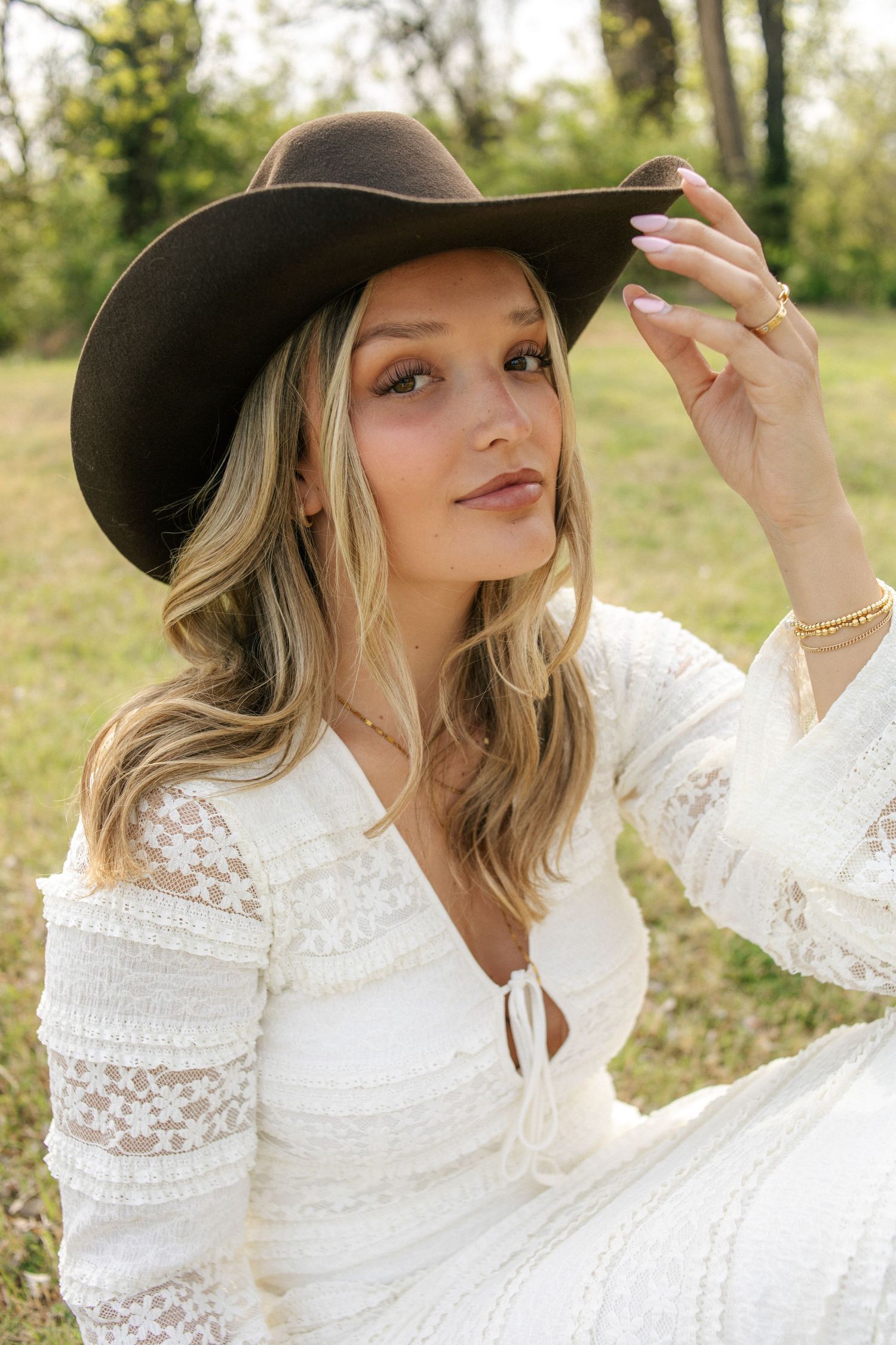 Blonde woman in white lace dress and brown cowboy hat sitting outdoors in a green field.