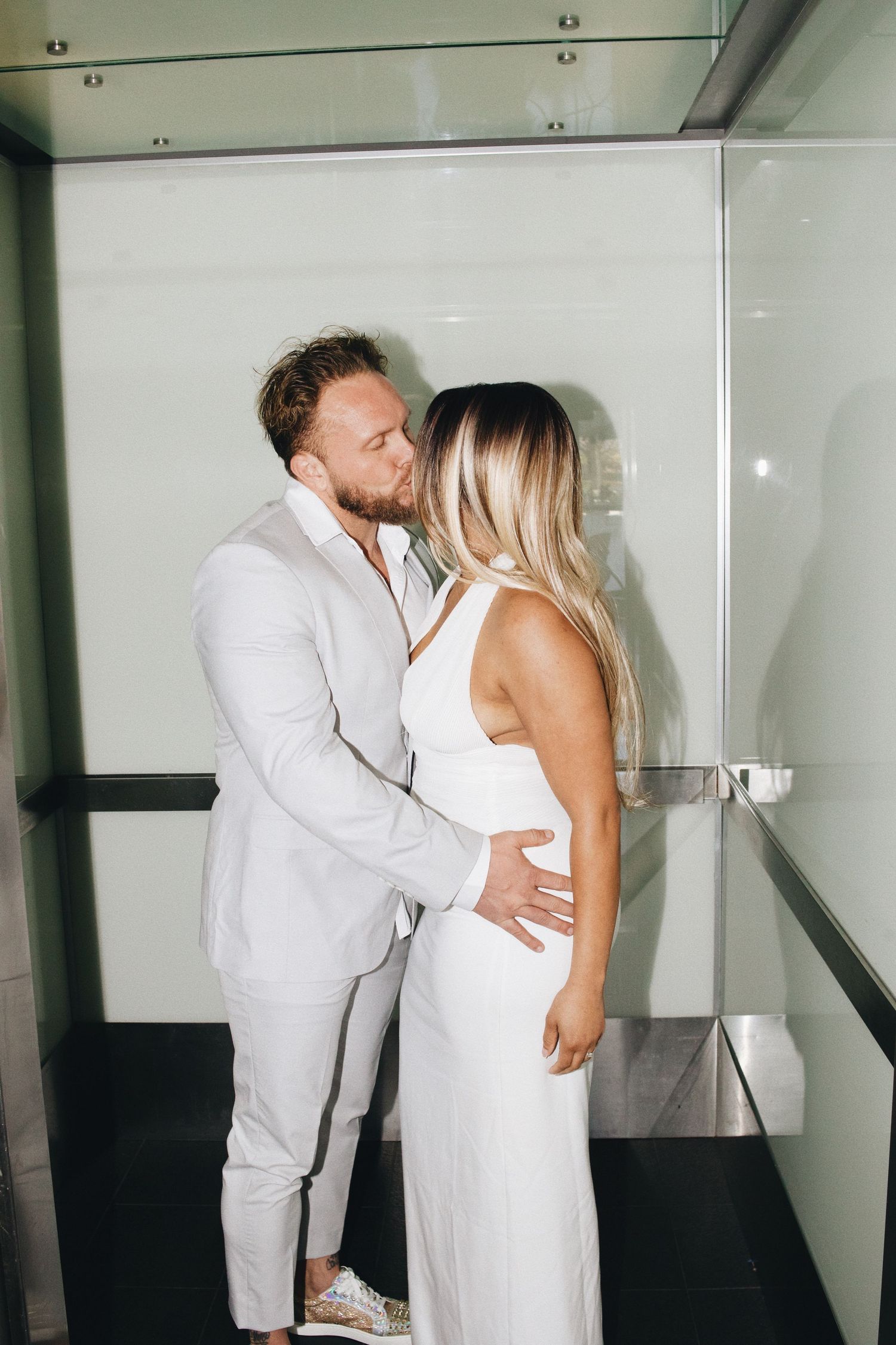 Couple in white outfits sharing a romantic kiss inside a modern glass elevator.