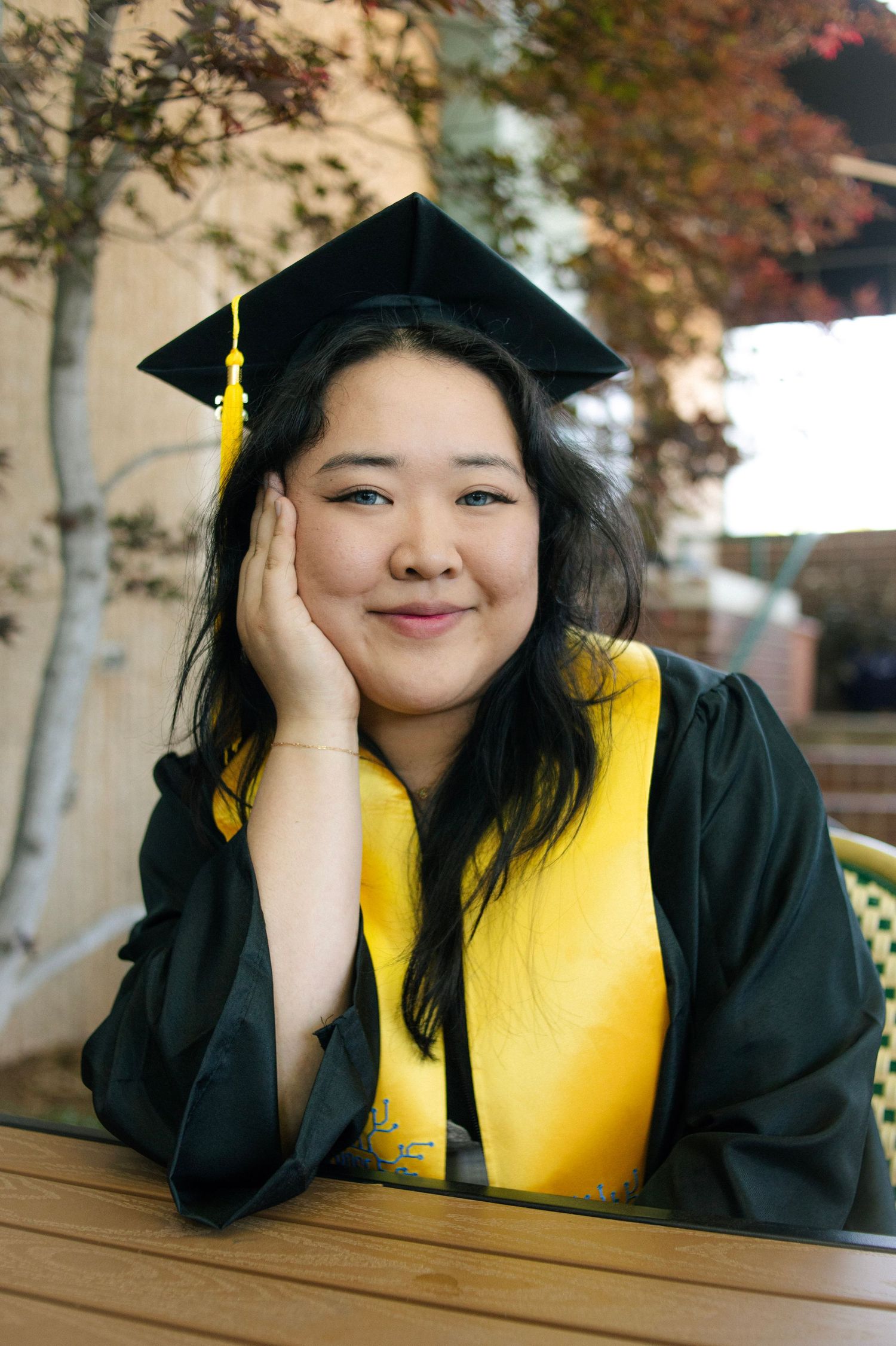 Smiling Asian woman in black graduation cap and gown with yellow stole posing at outdoor table.