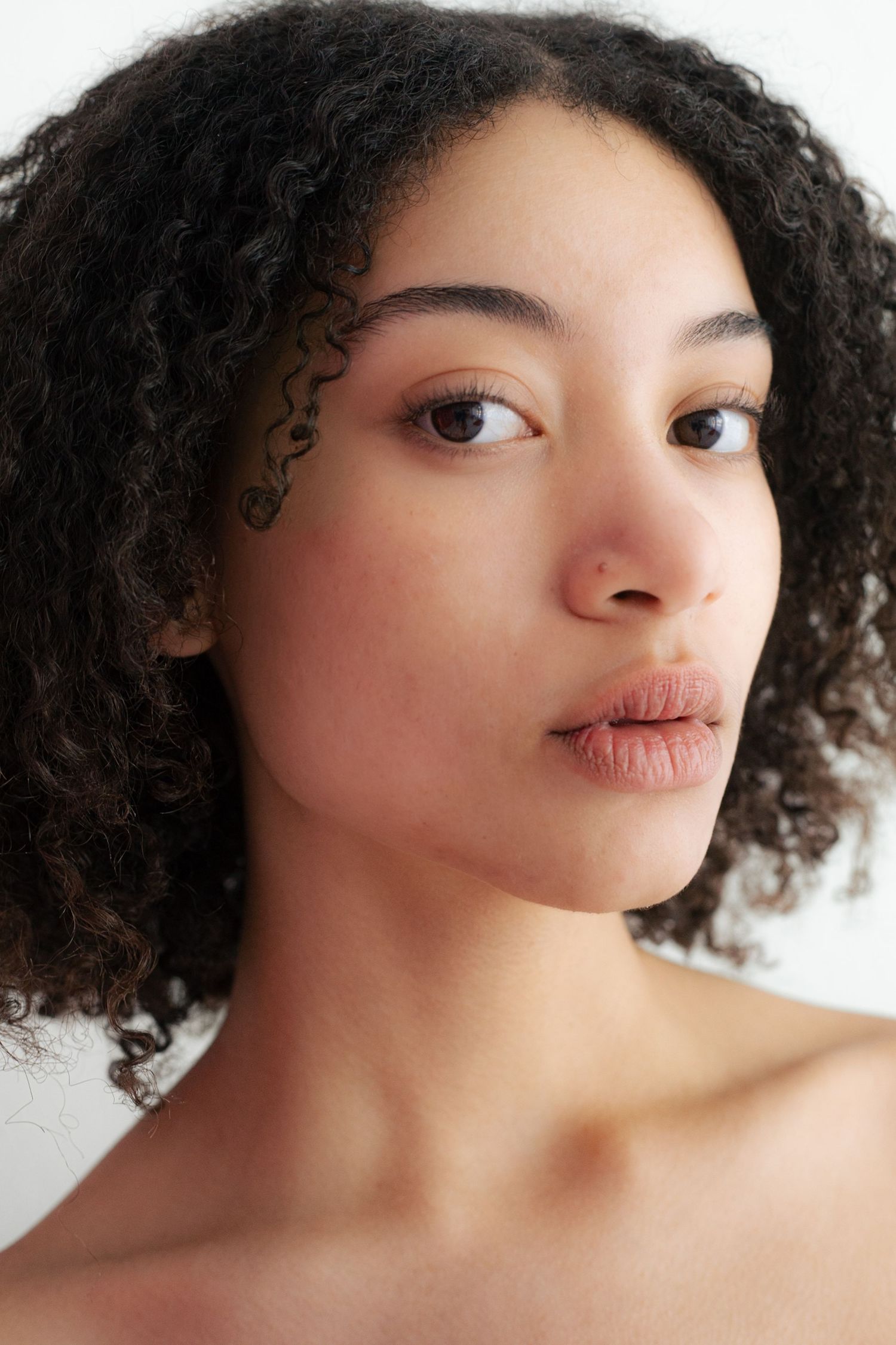 Close-up portrait of young woman with natural curly hair and bare shoulders on white background.