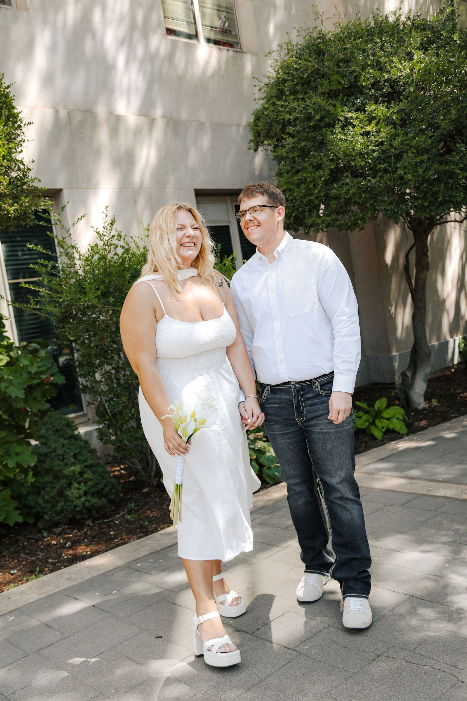 Happy couple in white outfits hold hands and walk together outdoors on a sunny urban pathway.