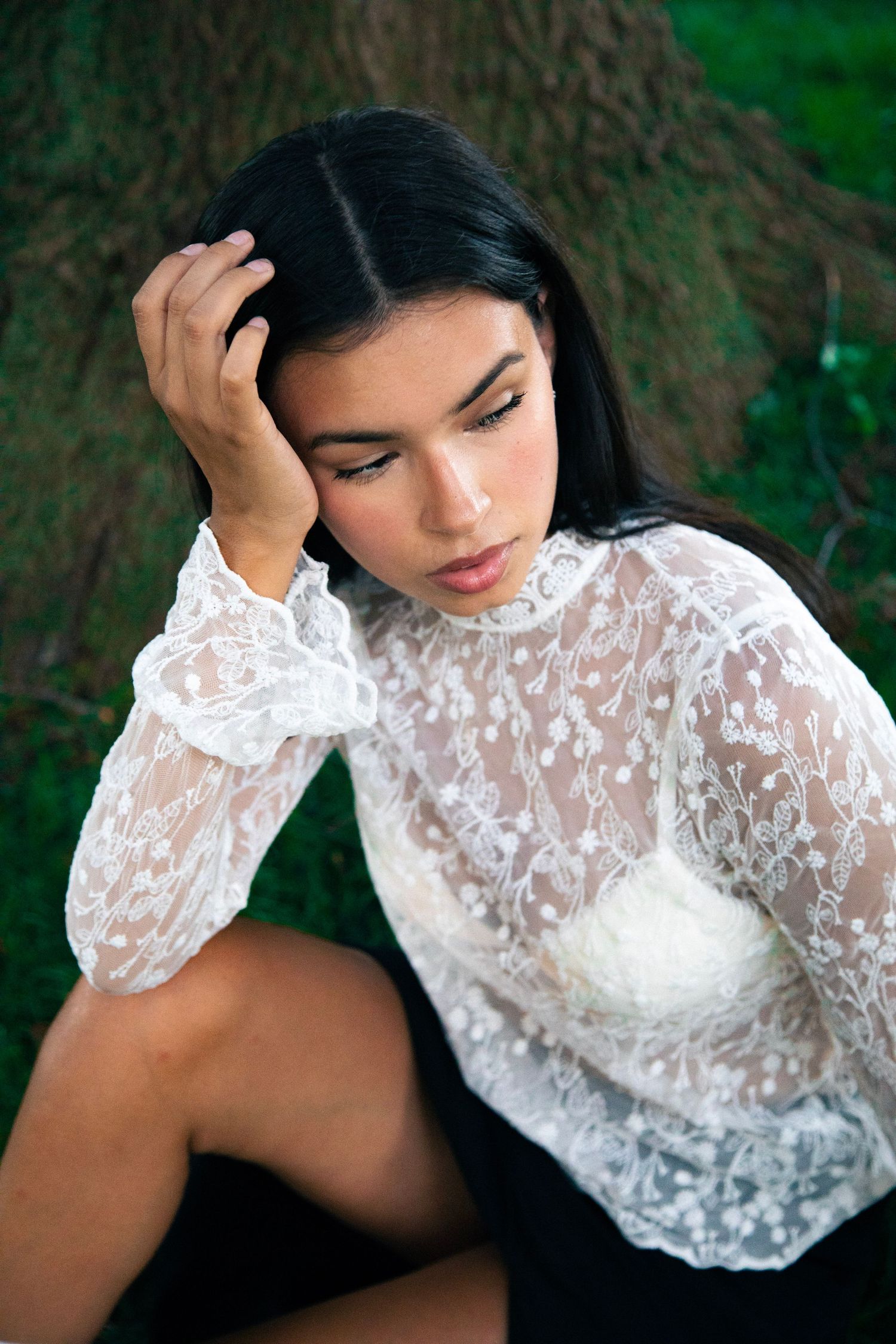 Dark-haired woman in white lace blouse sitting pensively against a tree outdoors.
