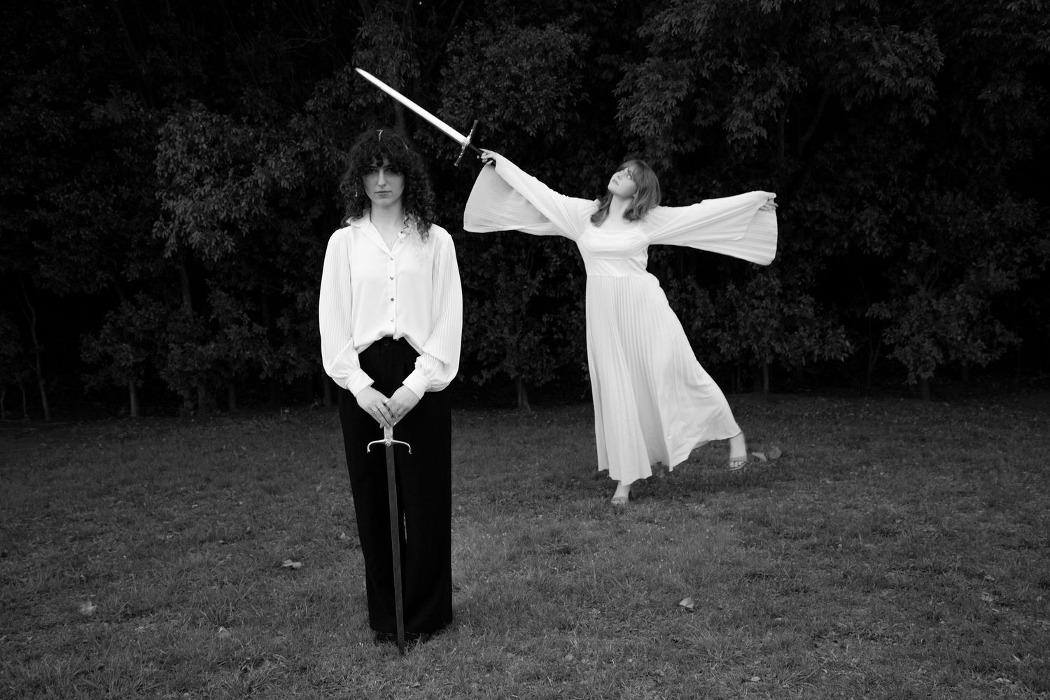 Black and white photo of two people in white outfits holding swords in a park.