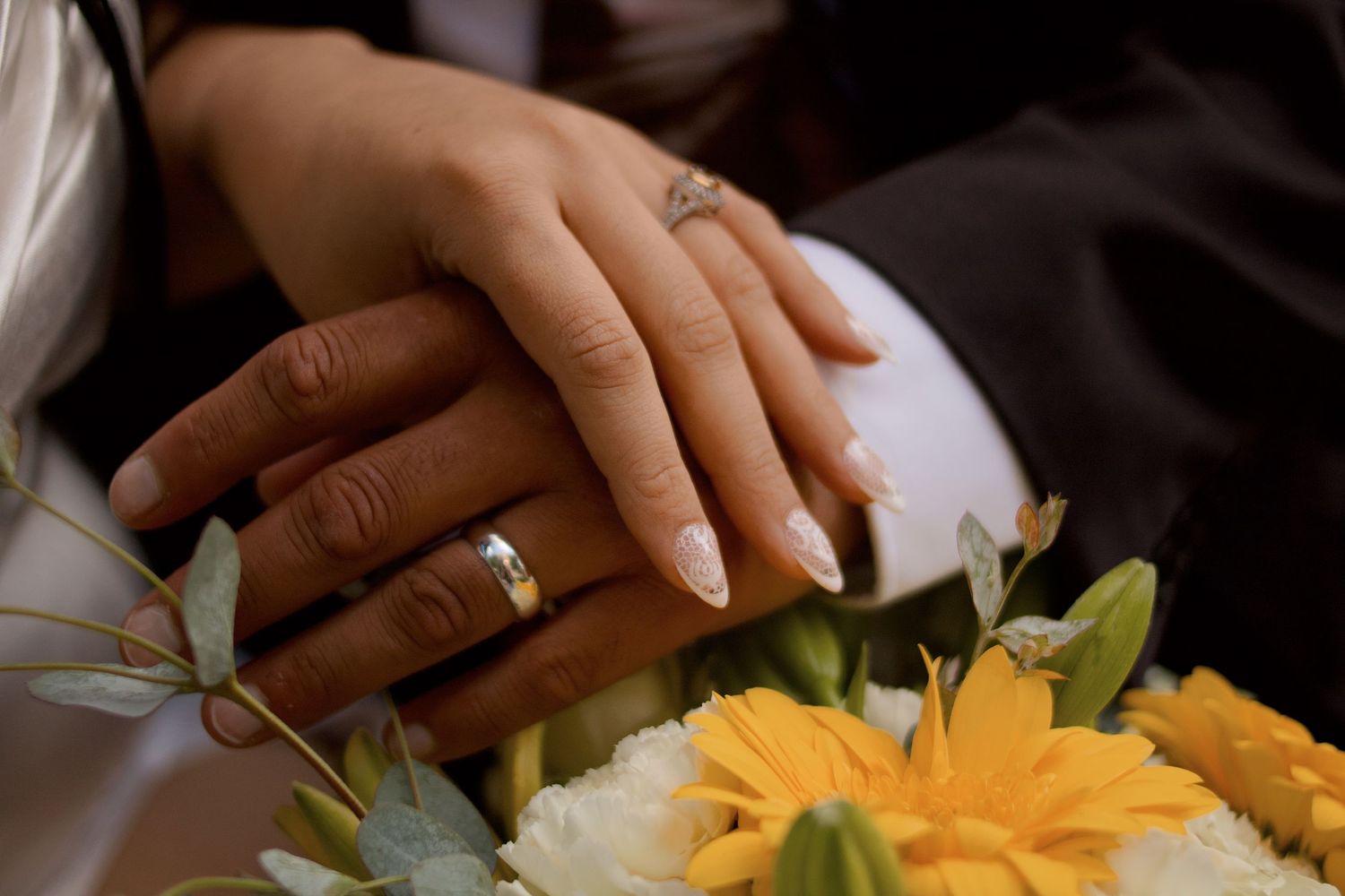 Close-up of newlywed hands with wedding rings resting on a bouquet of yellow and white flowers.