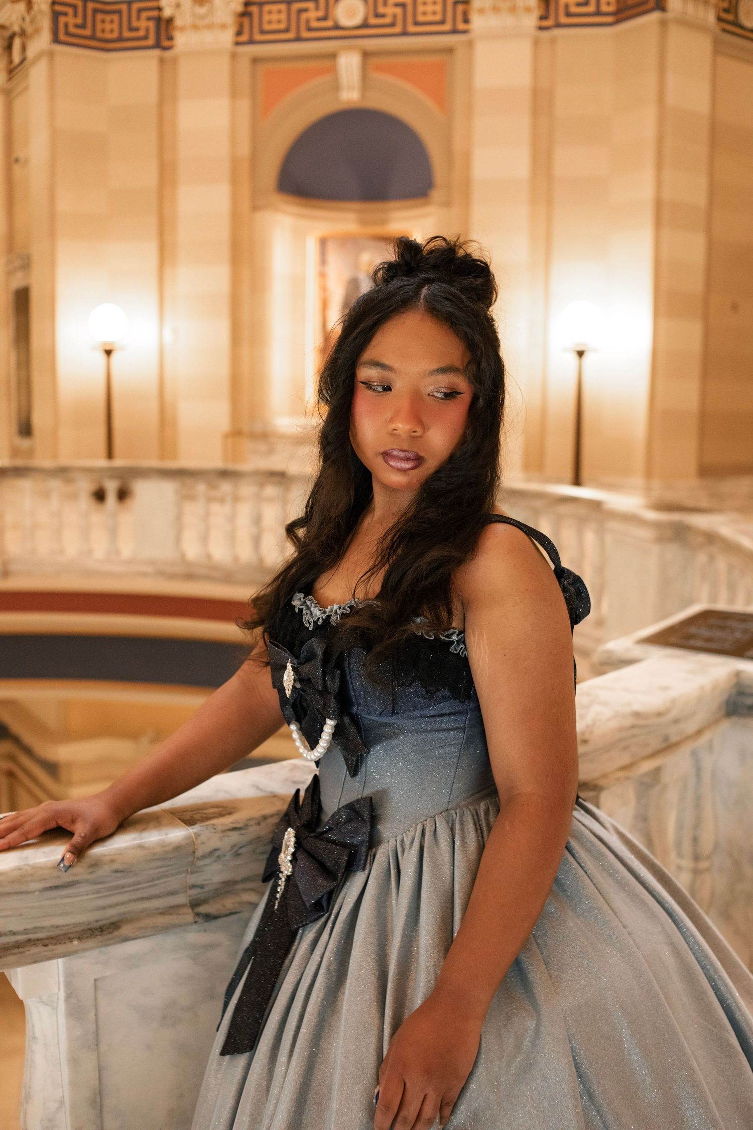Young woman in elegant blue-gray ball gown with black bows poses on marble staircase in ornate golden hall.