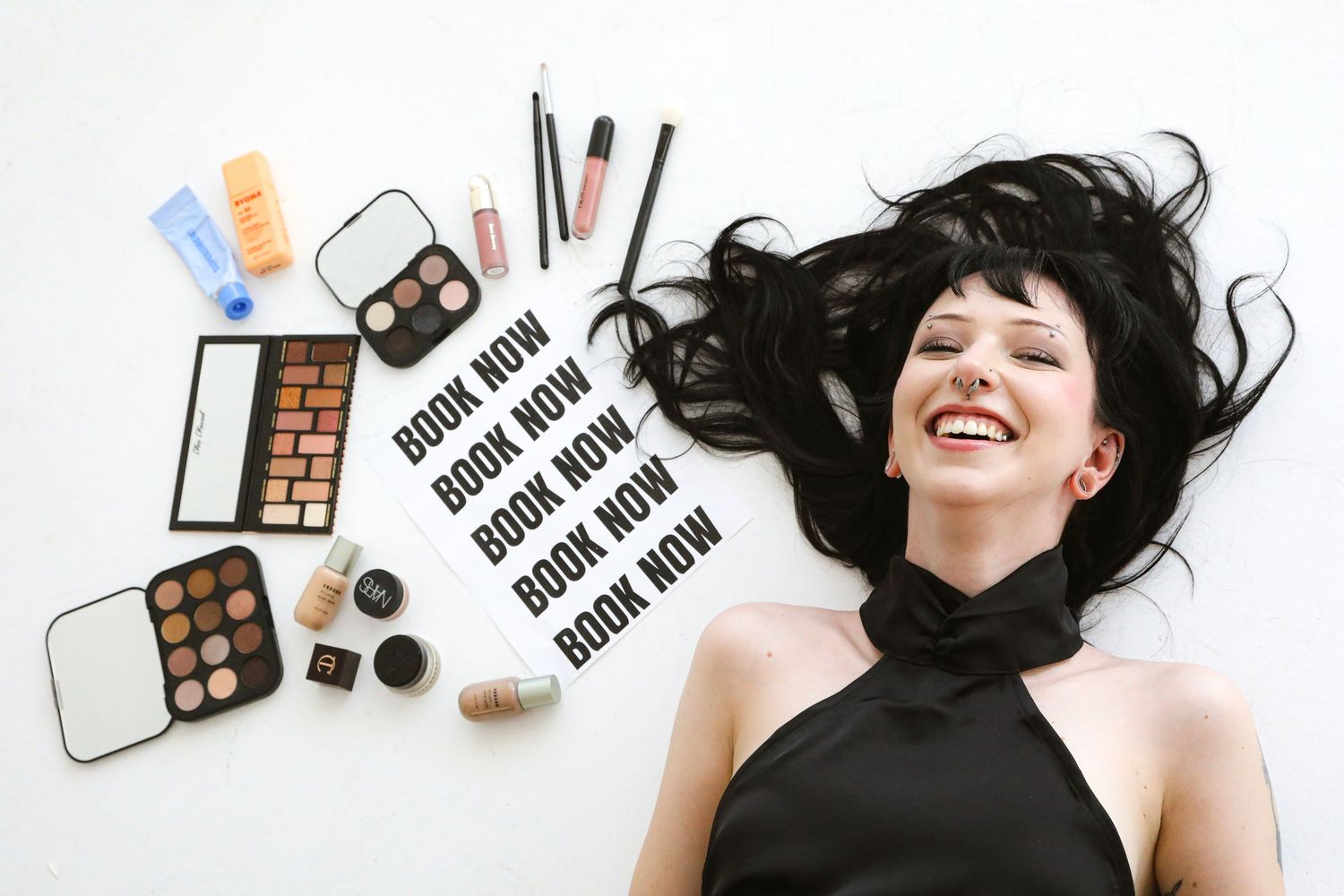 Woman lying among makeup products with book now text overlay, surrounded by eyeshadow palettes, lipsticks, and beauty essentials.