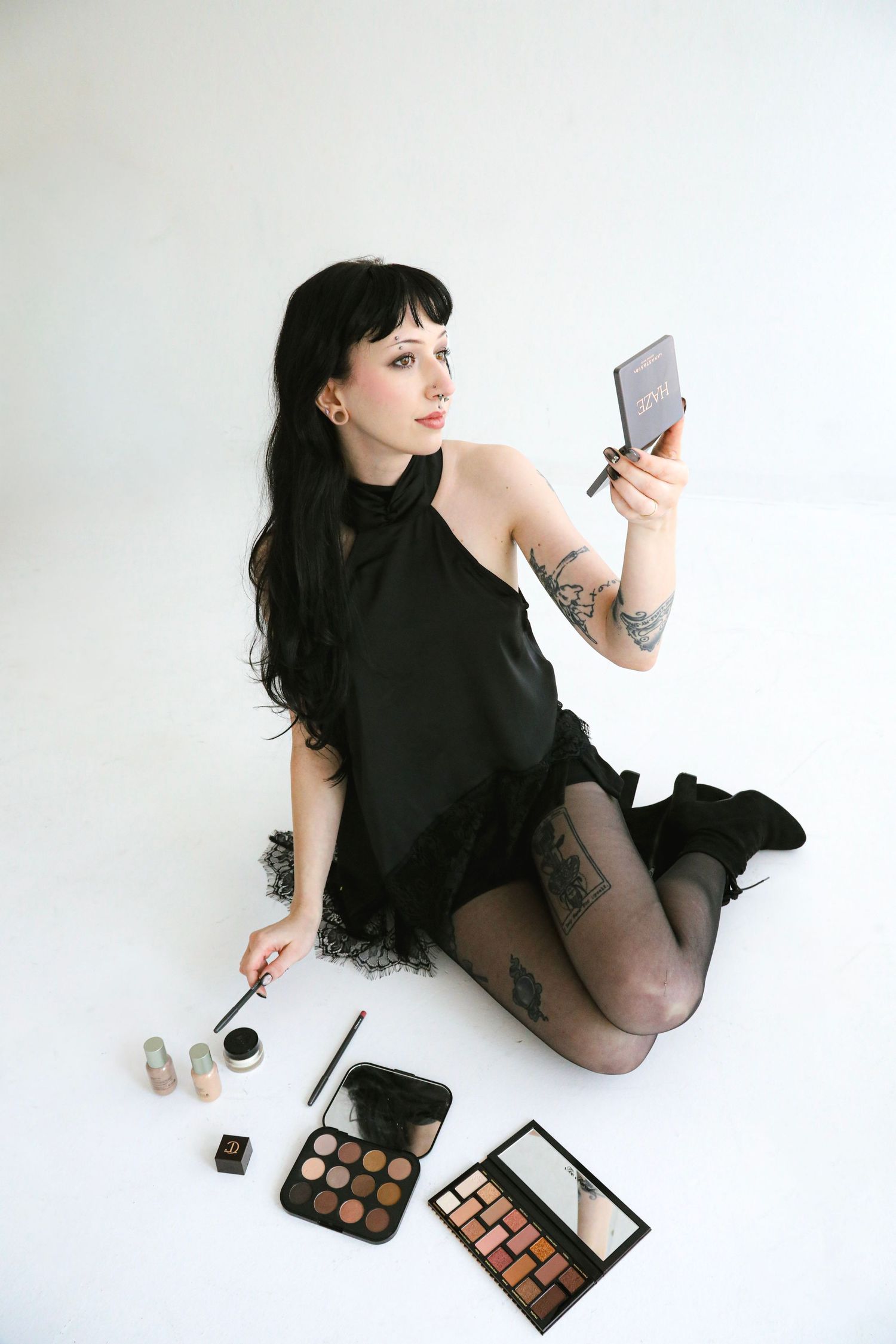 Woman in black dress sitting on white floor with makeup palettes, holding compact mirror and brush.