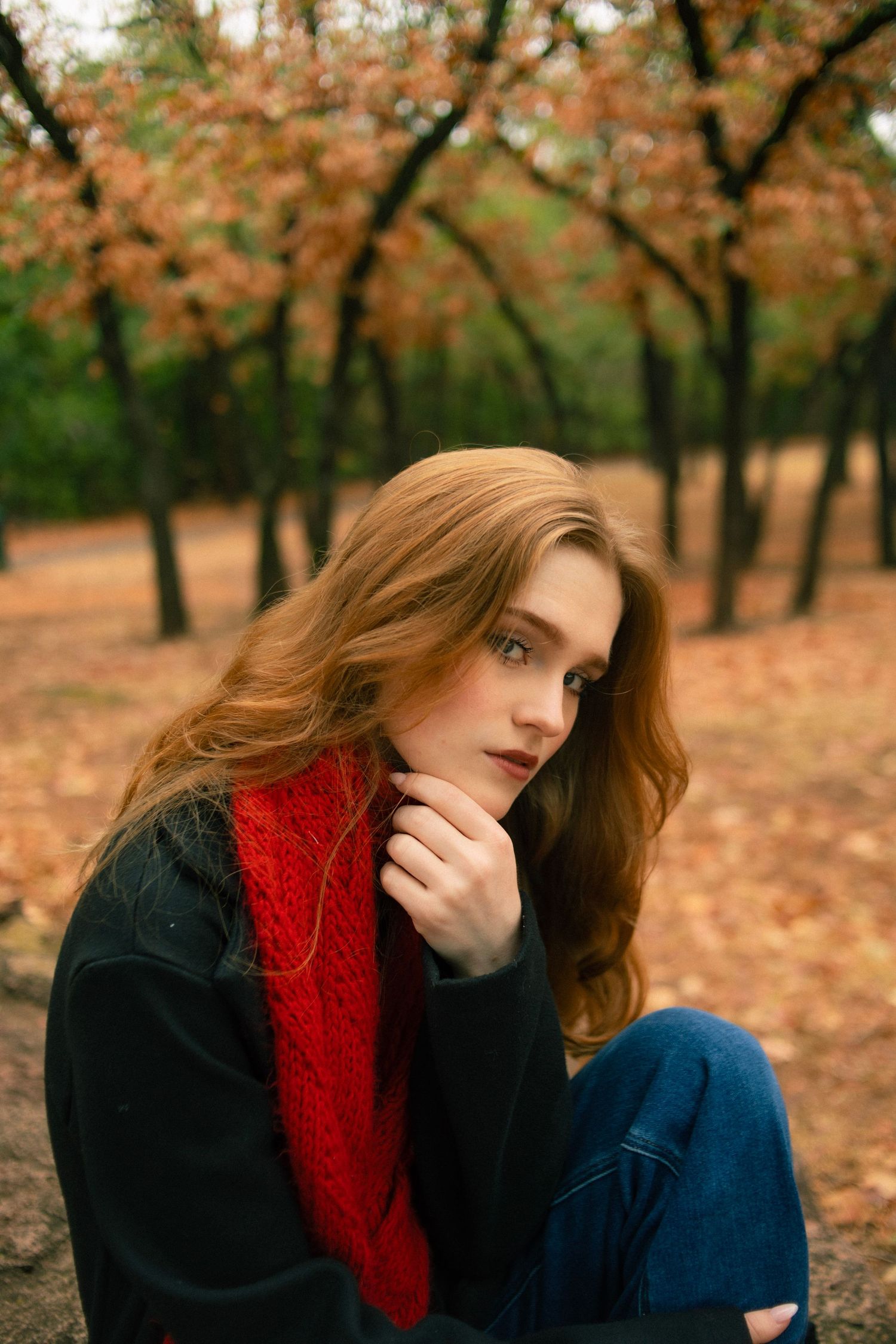 Young redhead woman in red scarf and black jacket sitting outdoors in autumn park with fallen leaves.