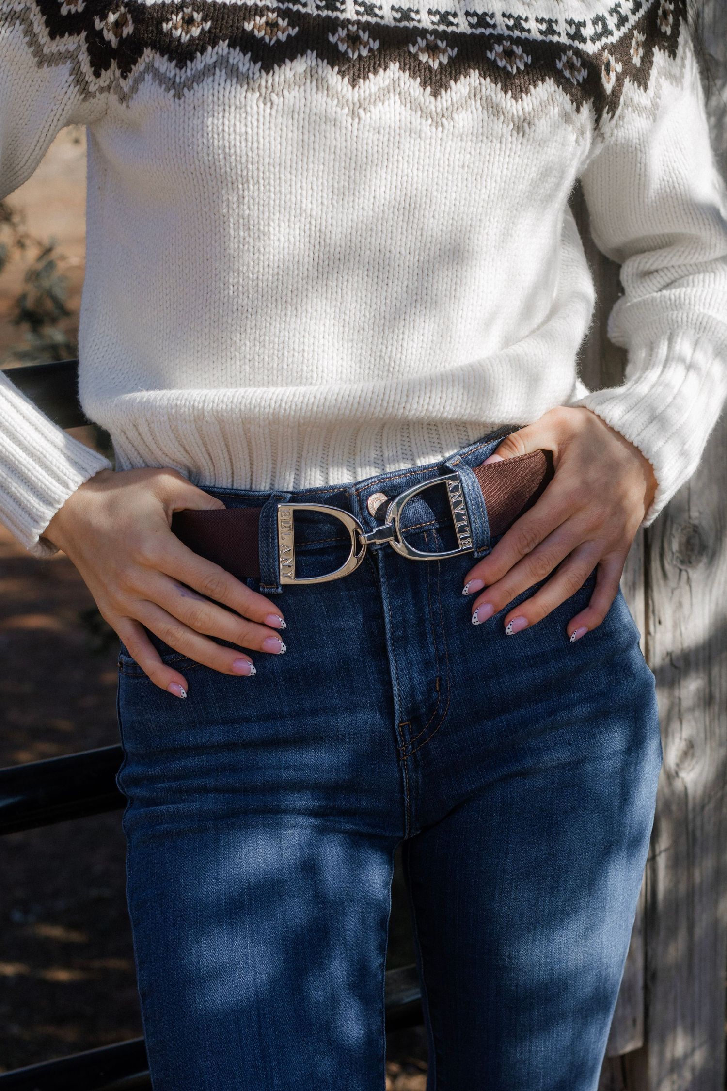 Close-up of woman wearing brown leather D-ring belt buckle with white knit sweater and blue jeans.