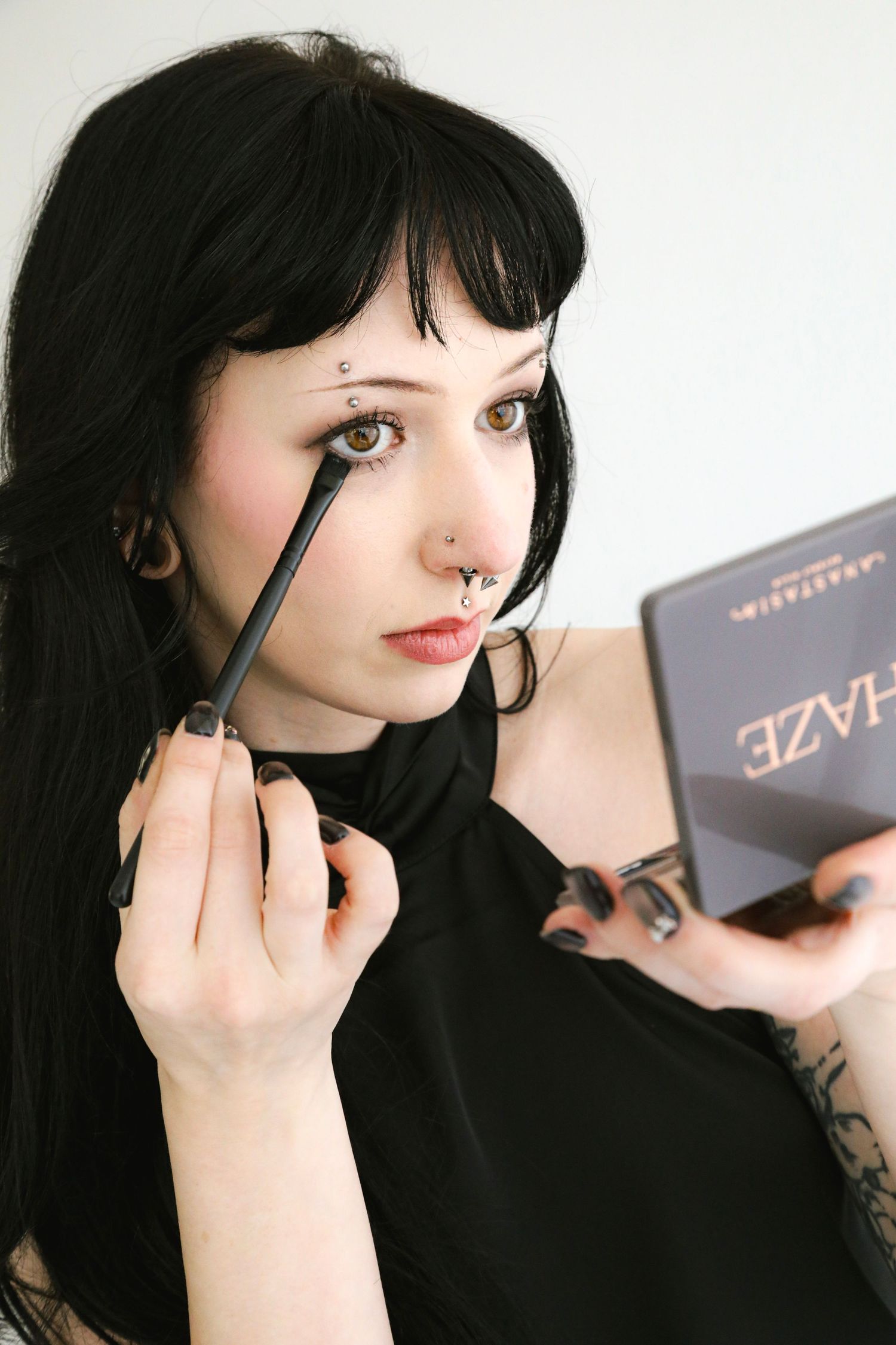 Young woman with black hair applying eye makeup using a brush, holding an Anastasia Beverly Hills Haze palette.