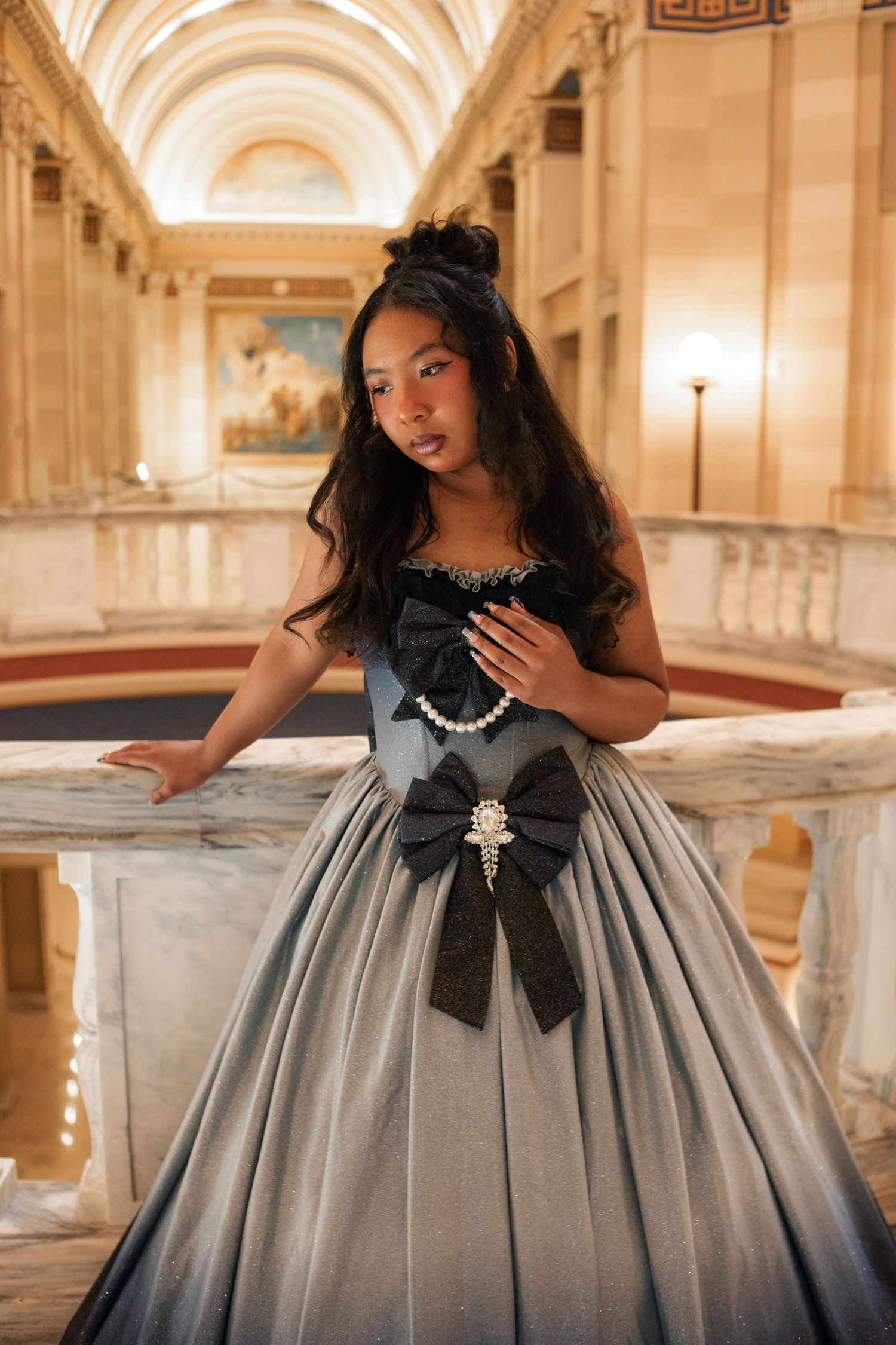 Young girl in elegant gray ballgown with black bow and pearl brooch poses on marble staircase in grand hall.