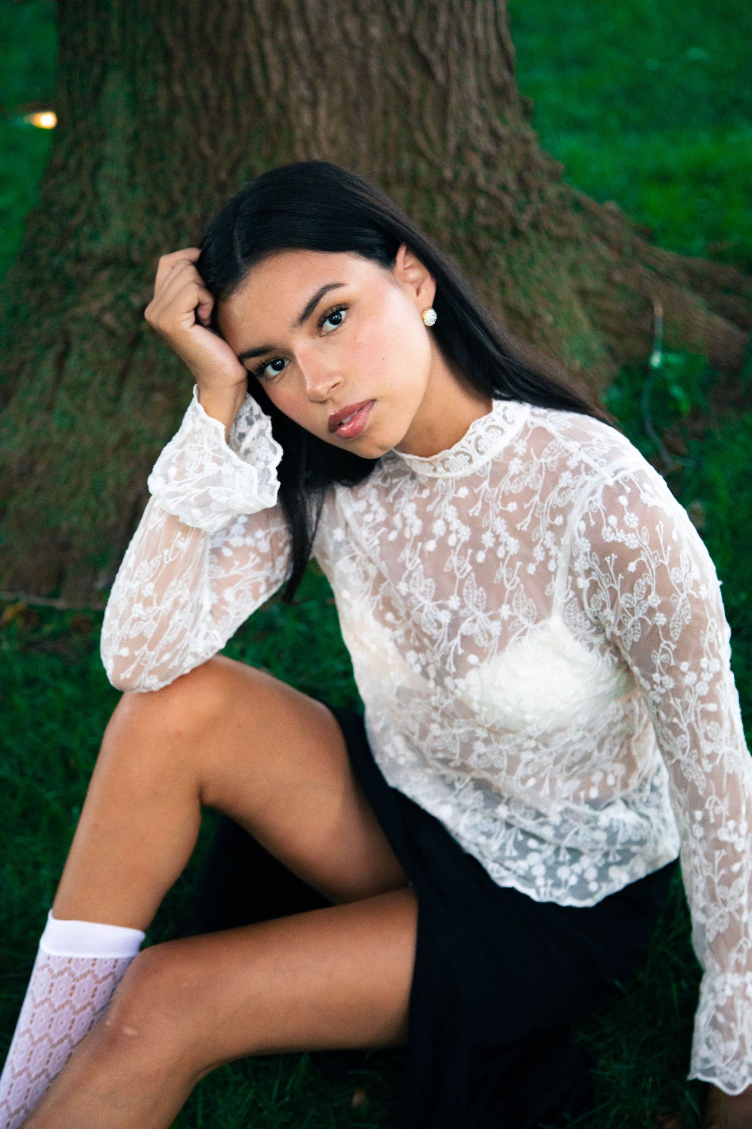 Young woman in white lace blouse and black skirt sitting against a tree wearing white knee socks.