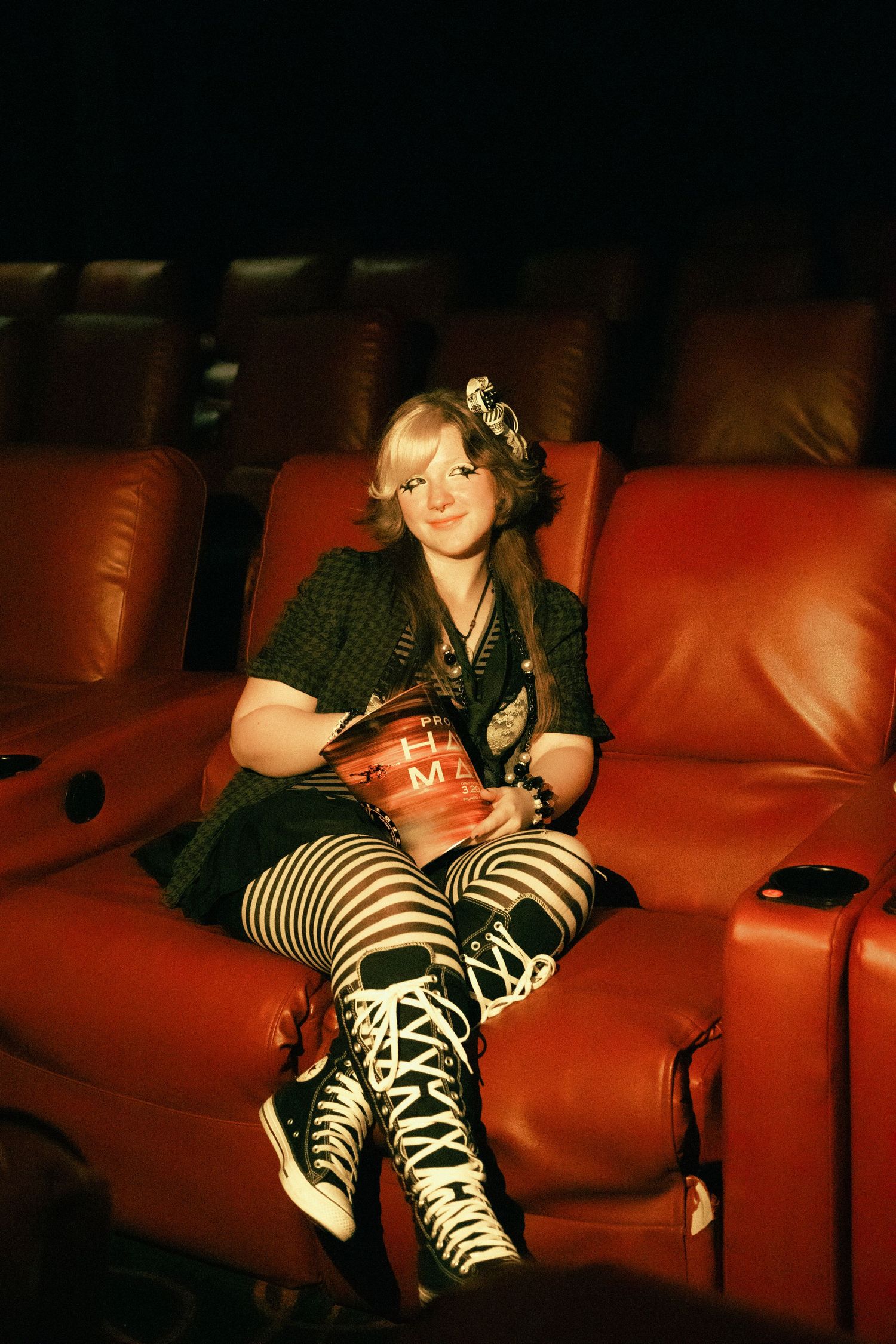 Young woman in striped tights and plaid outfit holding popcorn in a red leather cinema seat.