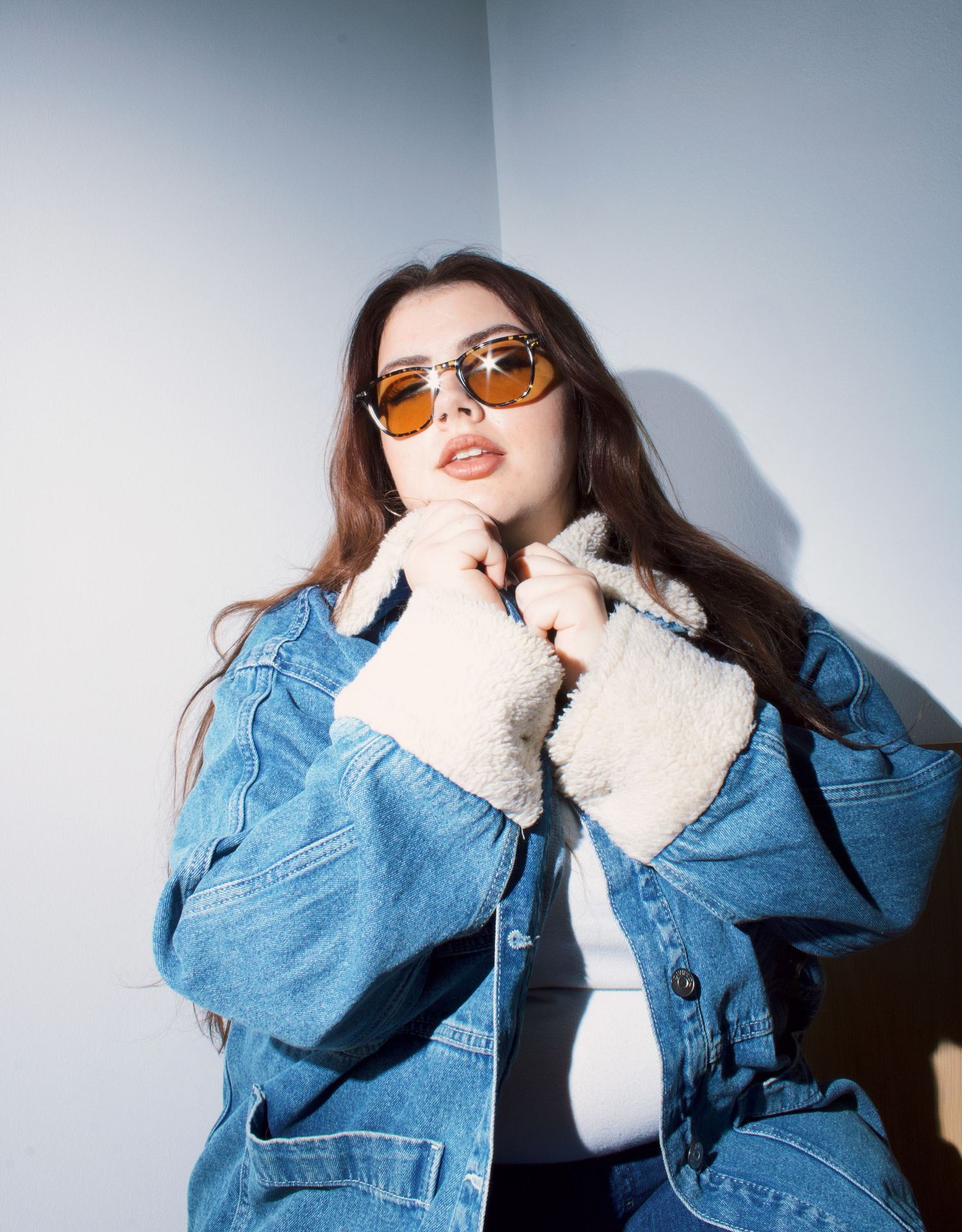 Brunette woman in oversized denim sherpa jacket and amber tinted sunglasses posing in corner.