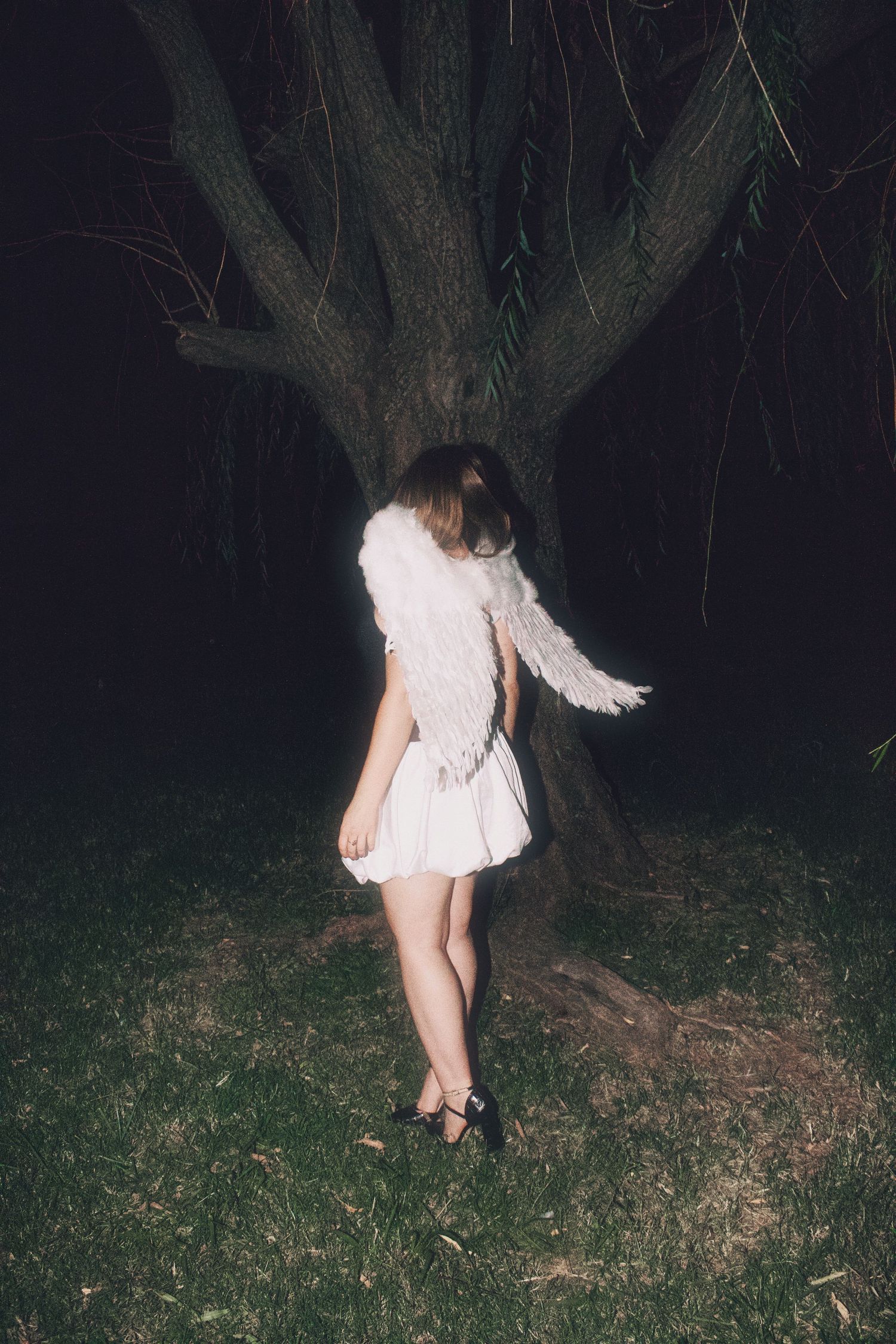 Girl in white mini dress with angel wings standing by large tree in dark night setting.