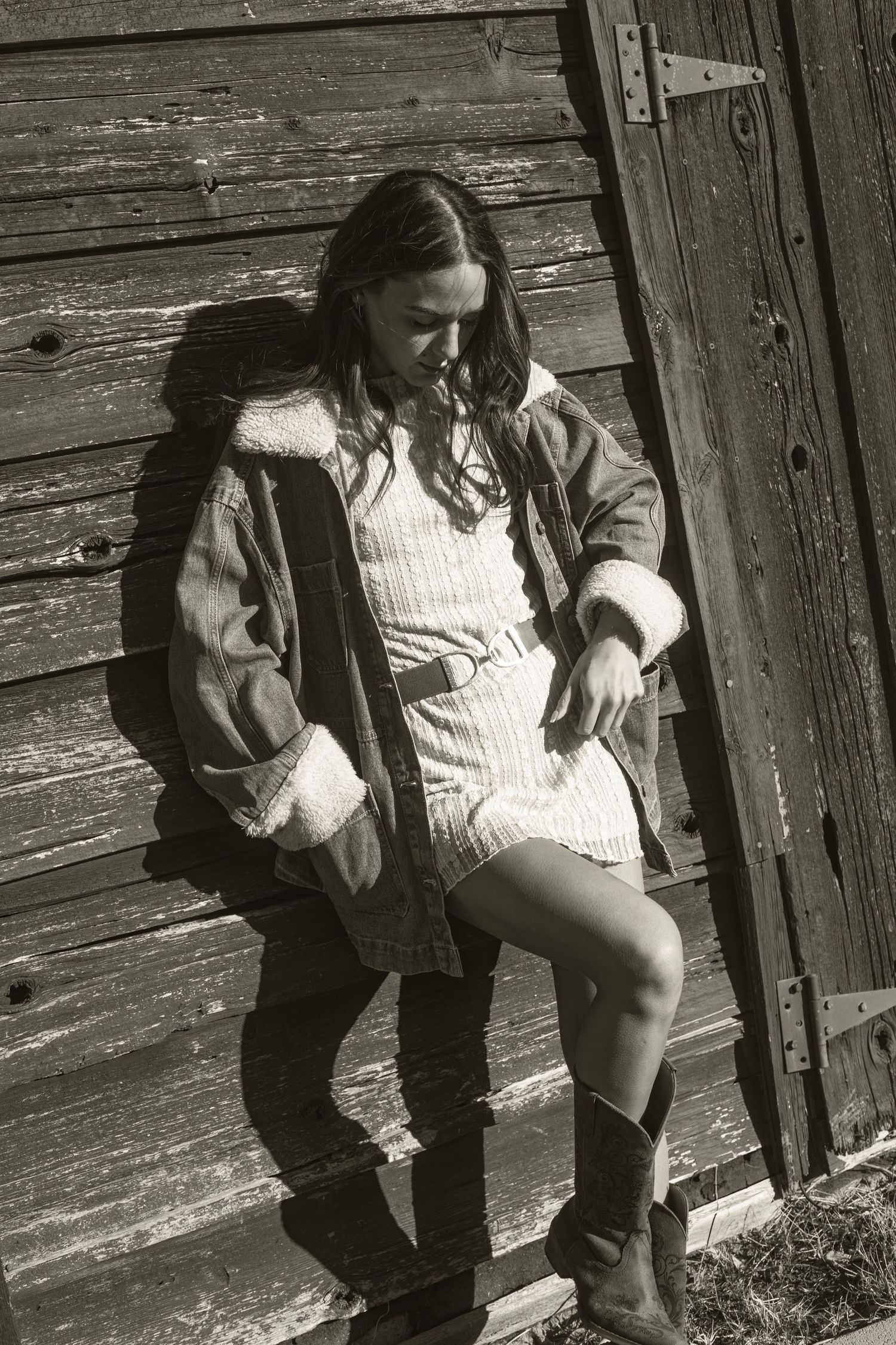 Black and white photo of woman in denim jacket and knit dress leaning against rustic barn door.