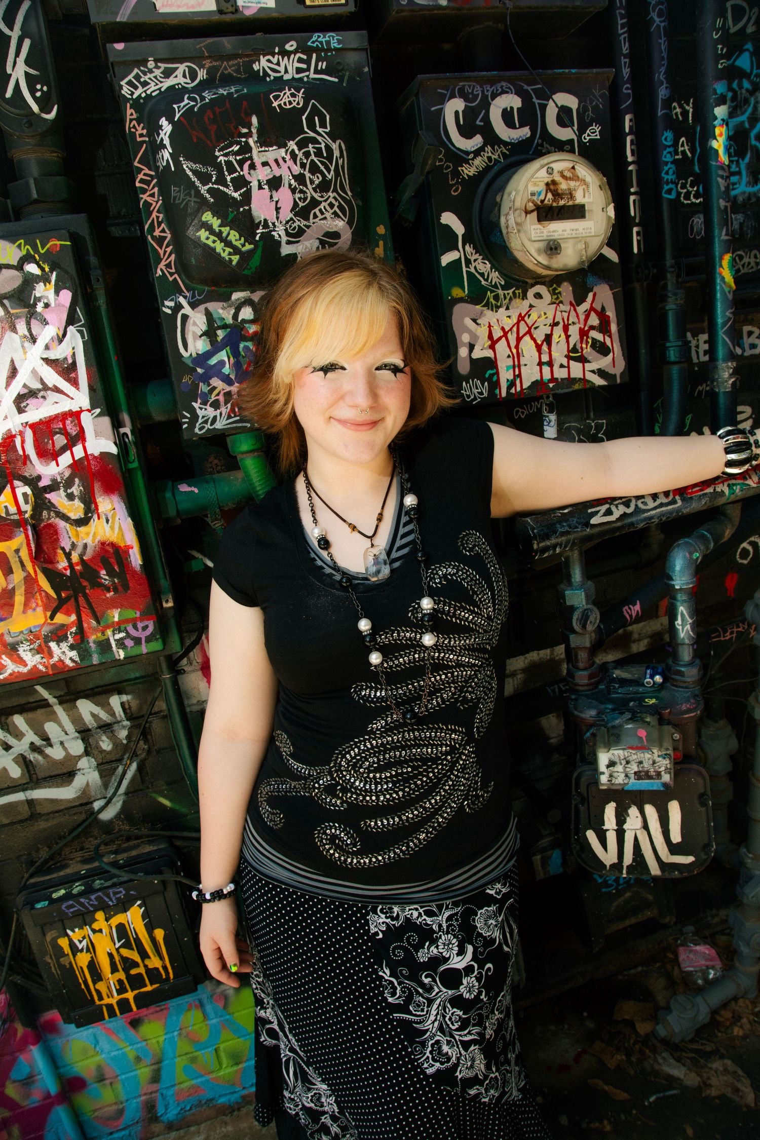 Young woman in black embroidered dress posing confidently against a graffiti-covered wall outdoors.