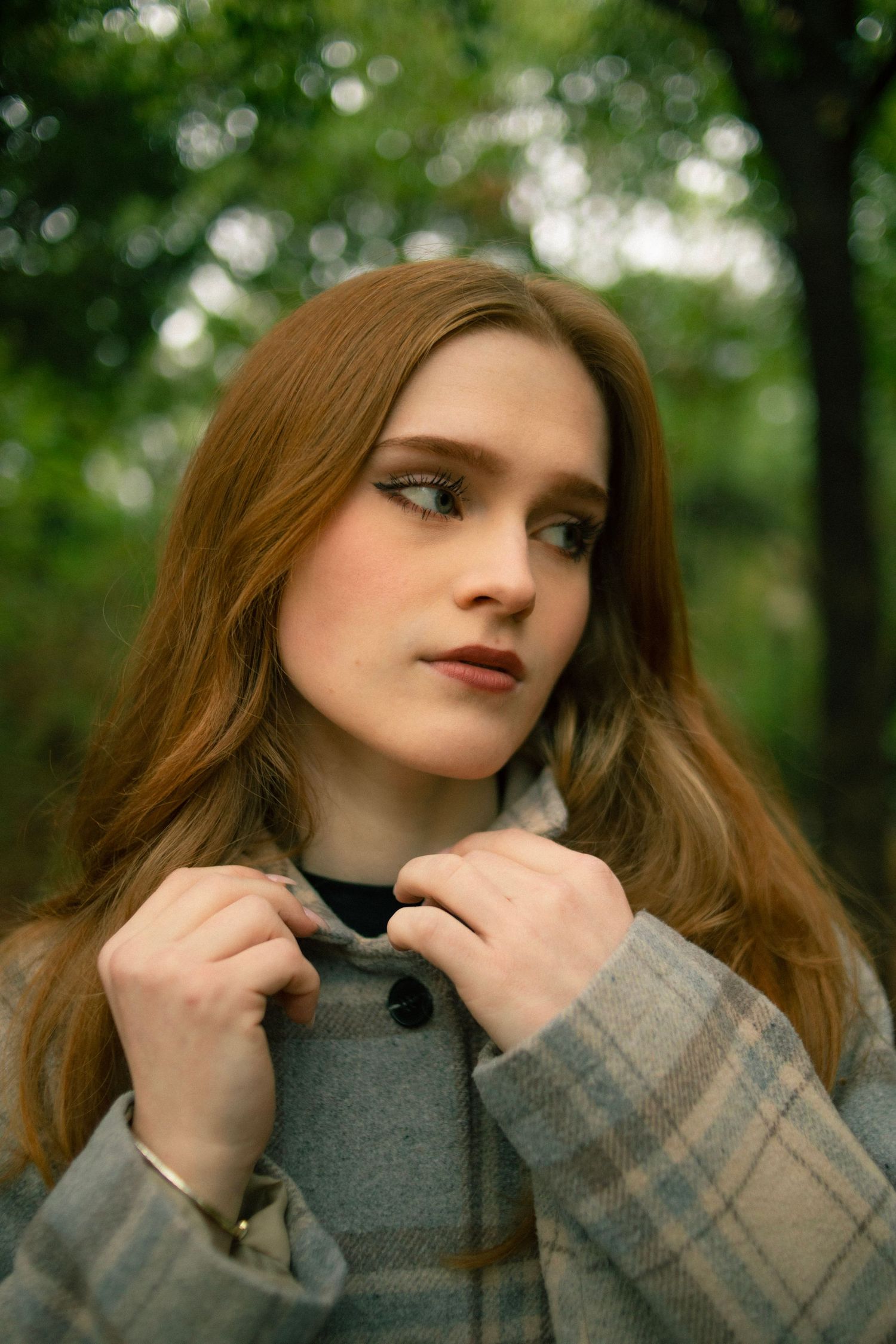Young woman with auburn hair in plaid coat poses outdoors among green trees in a portrait photo.