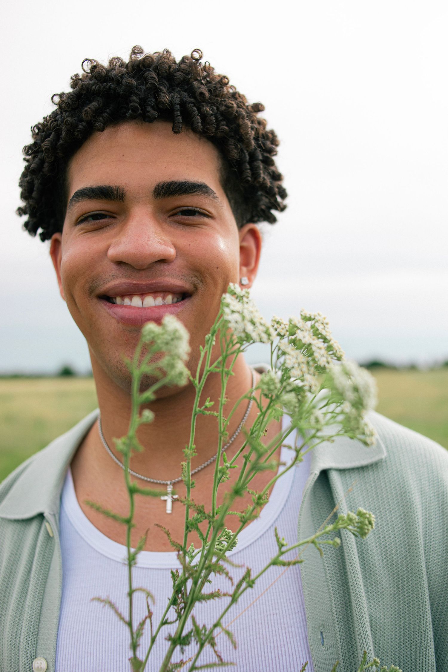 Smiling young man with curly hair holding white wildflowers in an open field wearing a cross necklace.