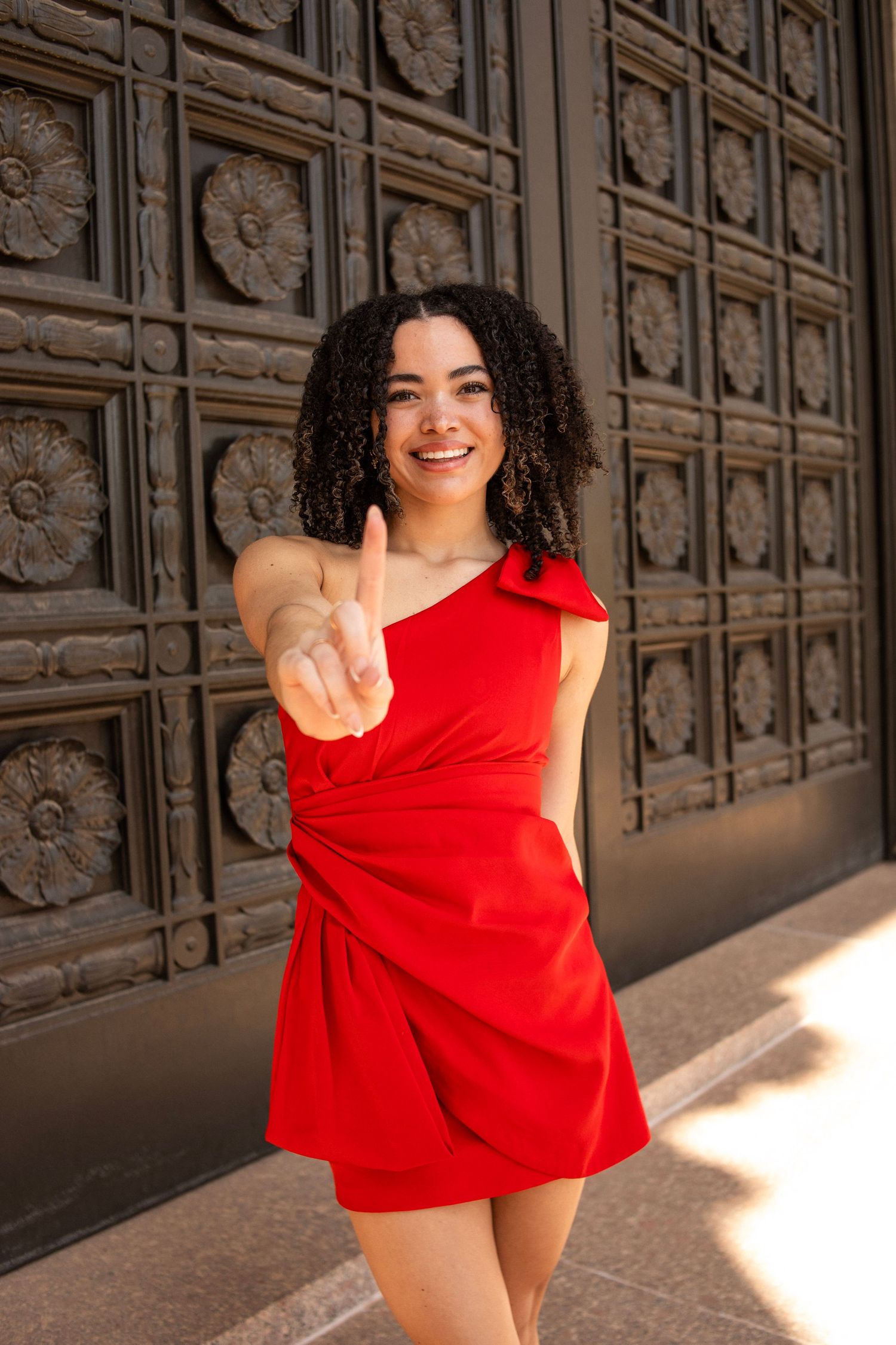 Smiling woman in red mini dress pointing finger toward camera outdoors.
