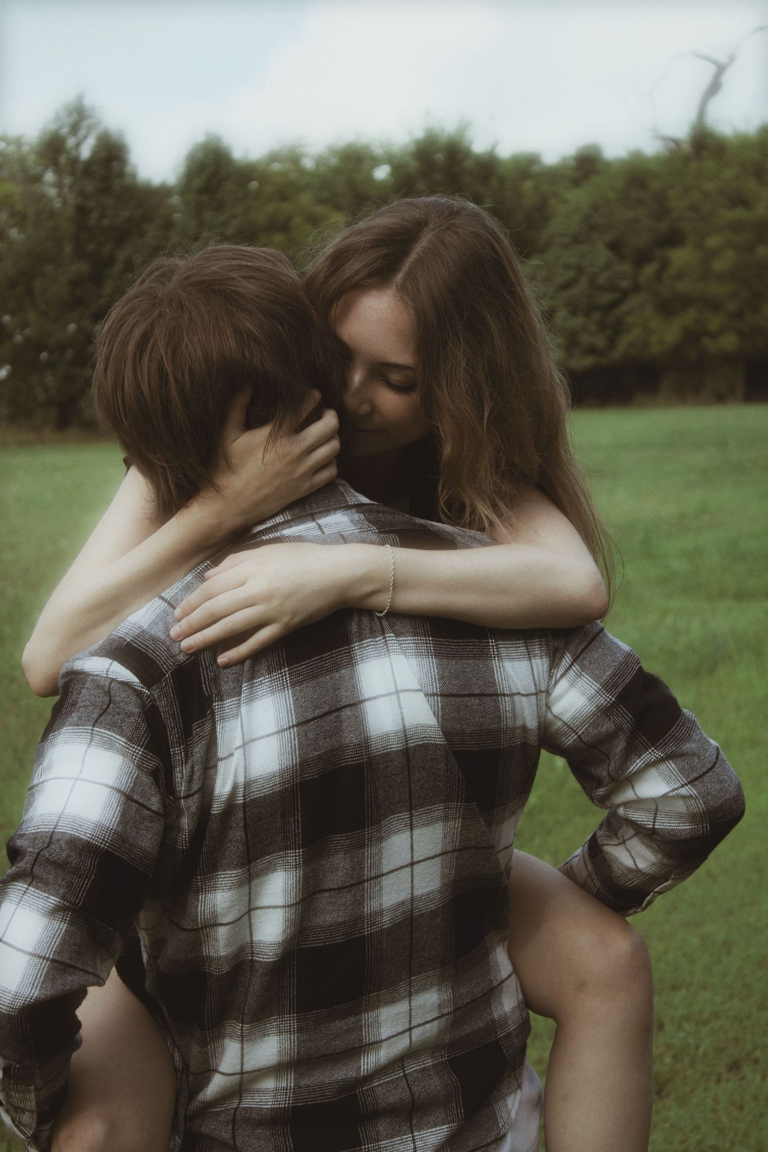 A couple shares an intimate embrace outdoors, with the man wearing a plaid shirt in a green field.