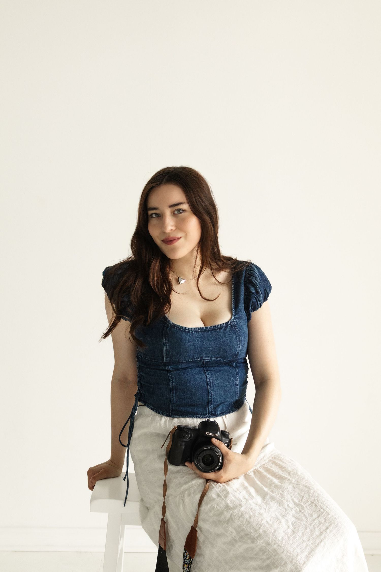 Brunette woman in denim top holds a camera while sitting against a white studio background.