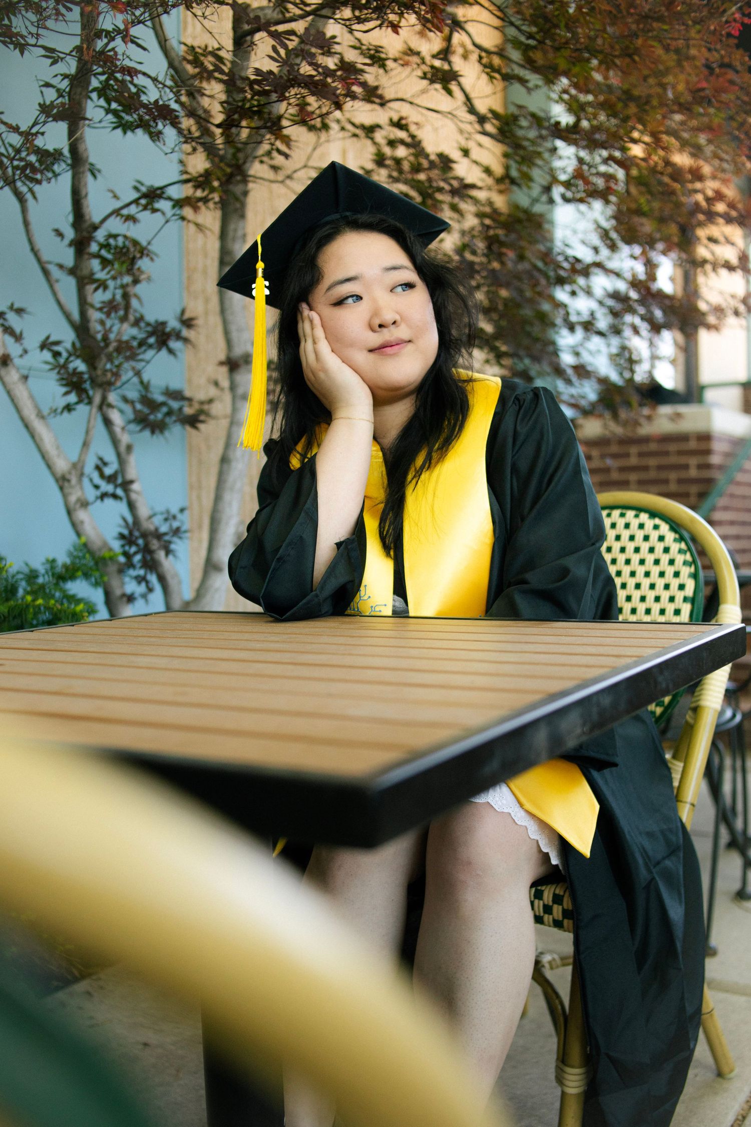 Young woman in green and gold graduation cap and gown sits thoughtfully at outdoor table with trees in background.