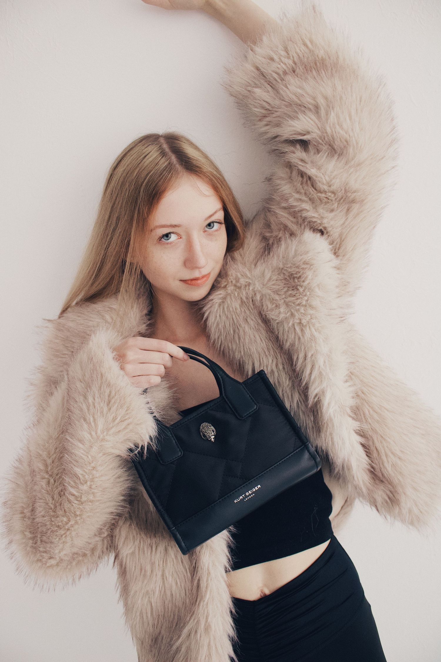 Young blonde woman in beige faux fur coat holding black Kurt Geiger handbag against white wall.