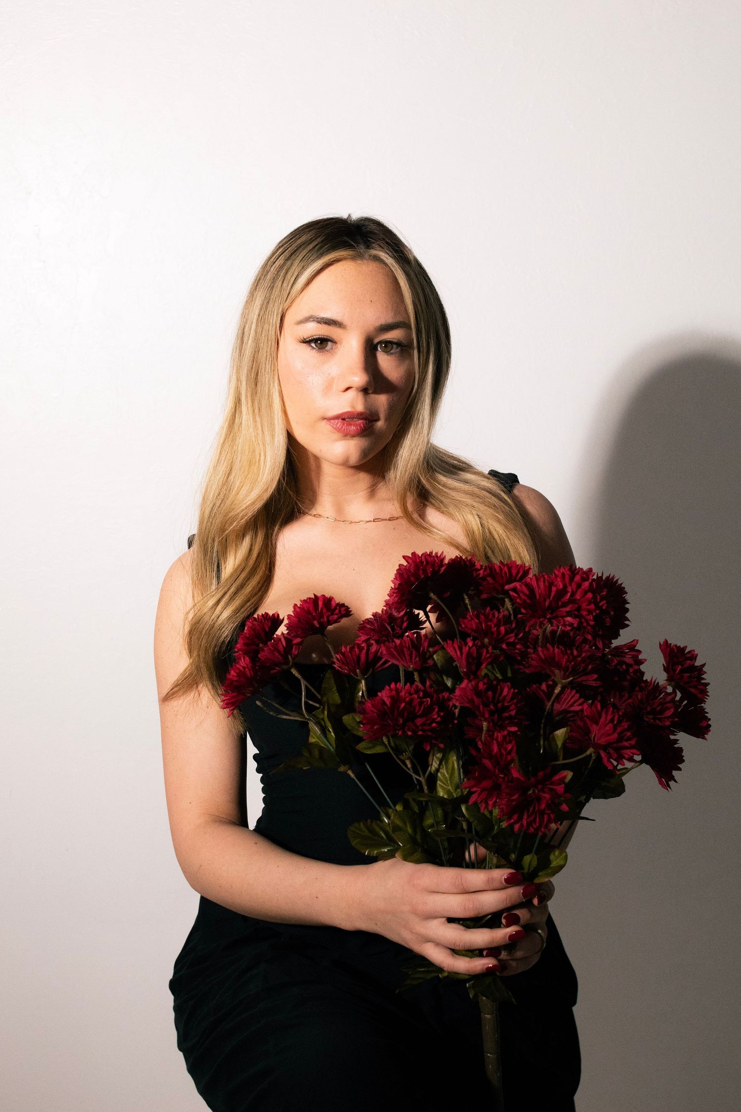 Woman in black strapless dress holding bouquet of dark red chrysanthemums against white background.