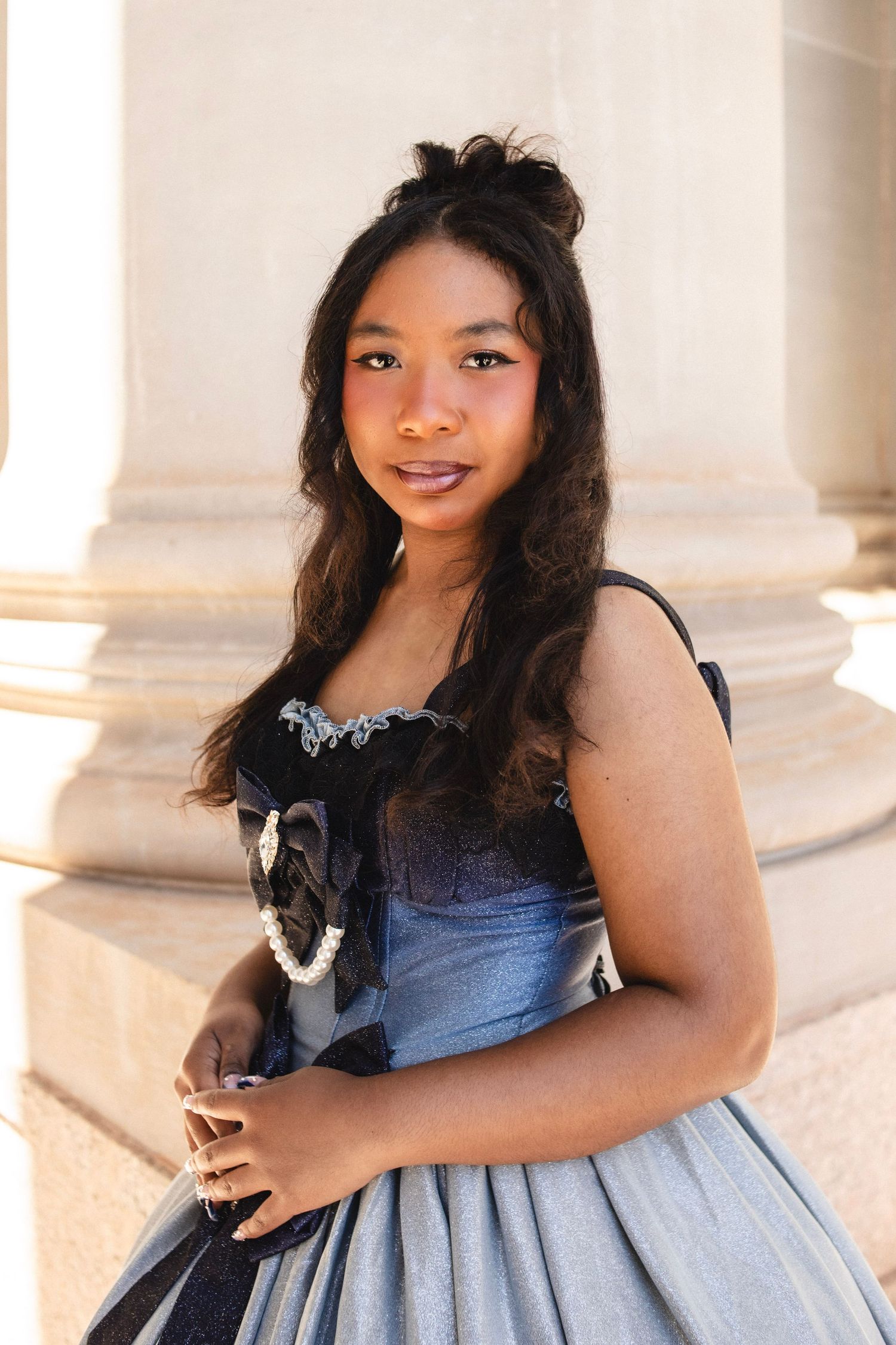 Young woman in vintage blue and black gown posing beside stone columns outdoors.