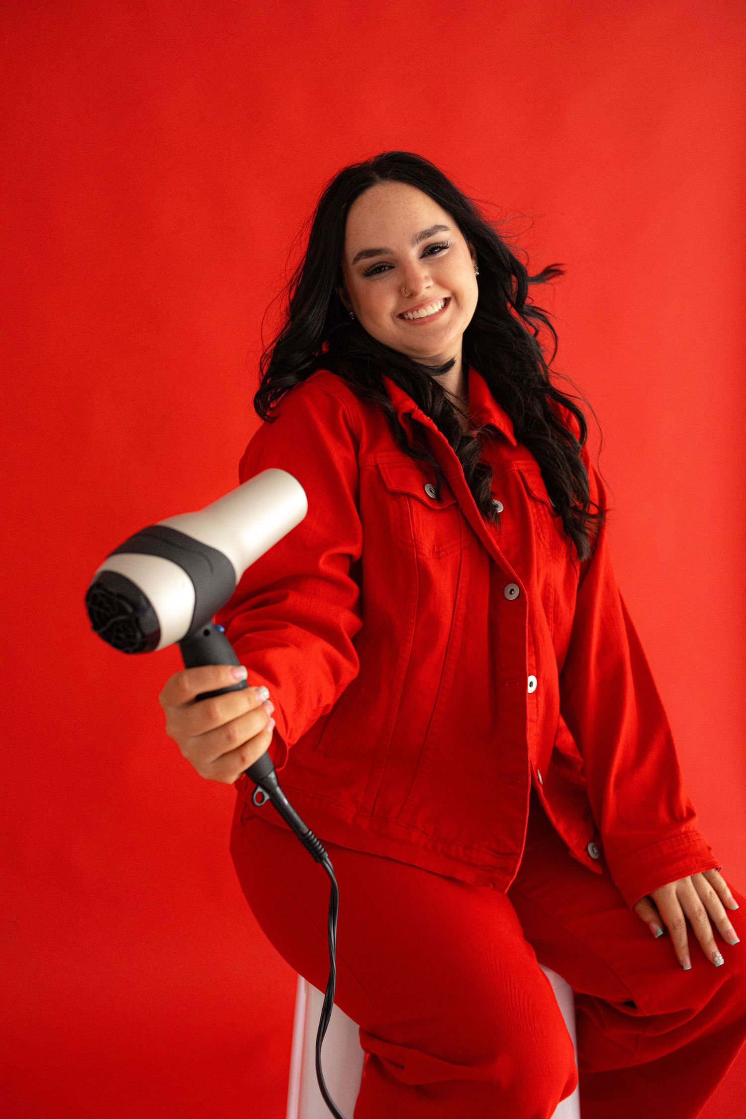 Smiling woman in red outfit holding hair dryer on bold red studio background.