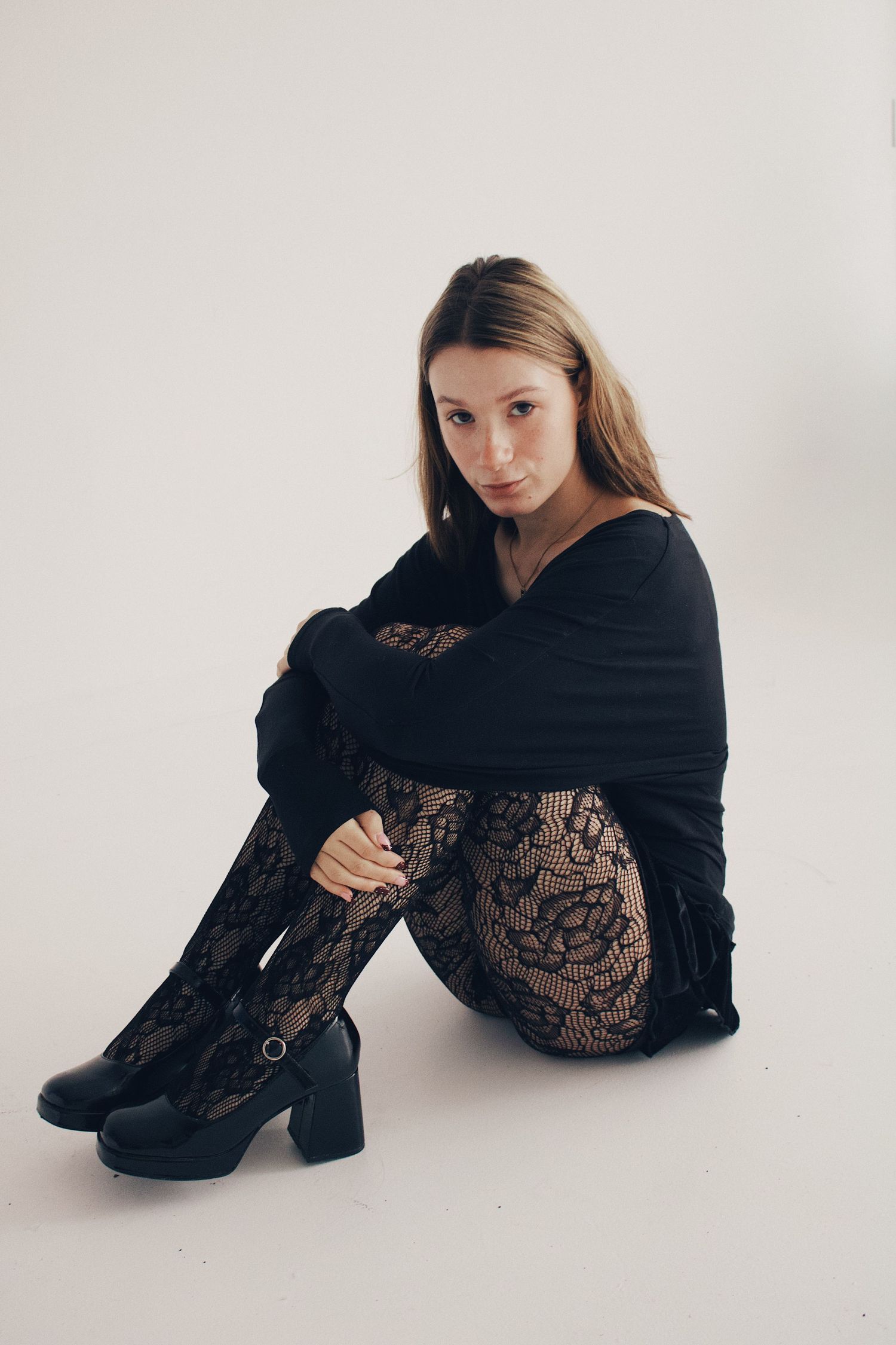 Young woman in black lace bodysuit and heeled boots sitting on white studio floor.