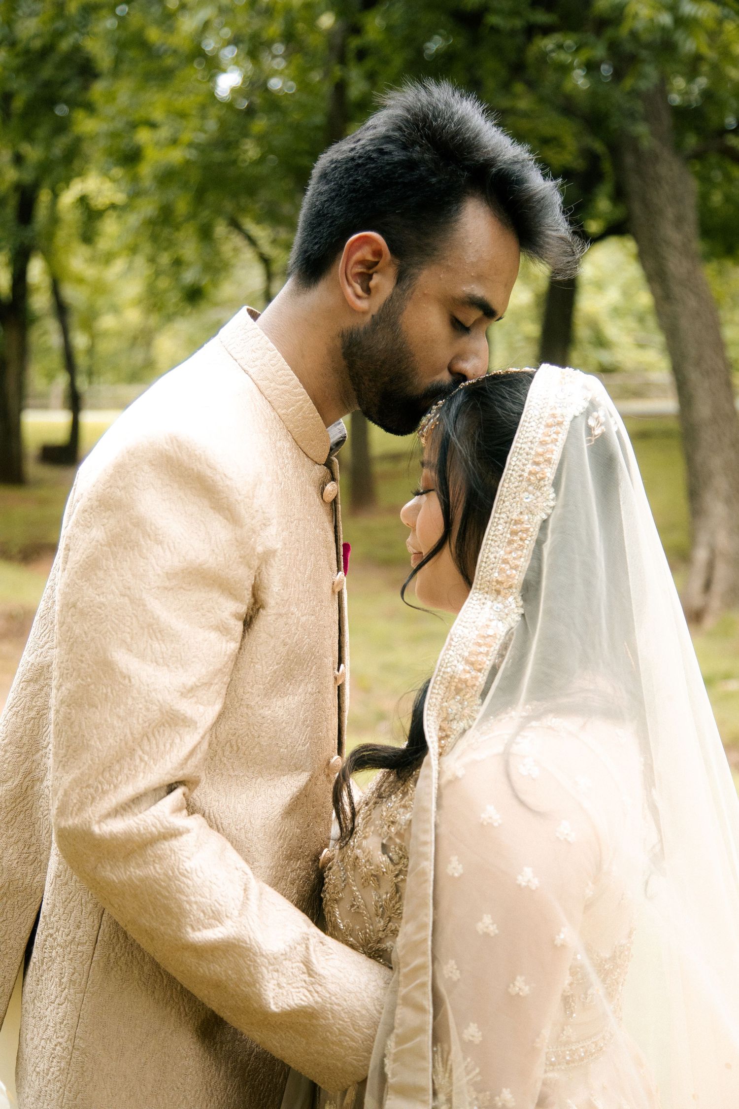 Groom in cream sherwani gently kissing bride's forehead while she wears an embroidered veil in a green park.