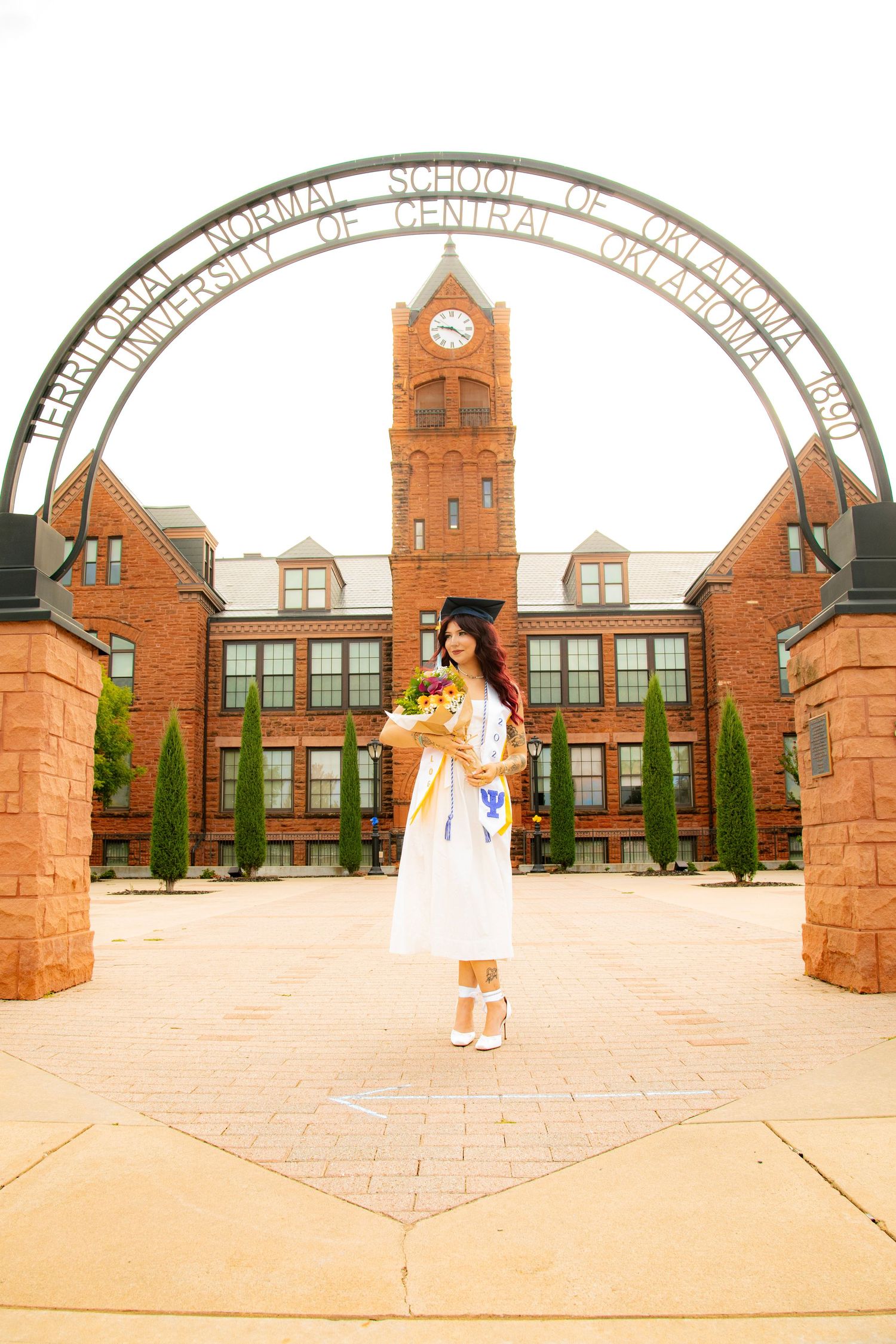 Graduate in white cap and gown holds flowers under University of Central Oklahoma arch on campus.