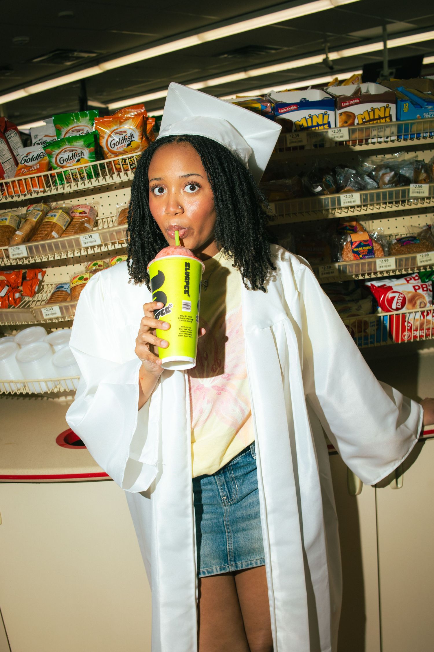 Graduate in white cap and gown sipping a large drink at a convenience store snack aisle.