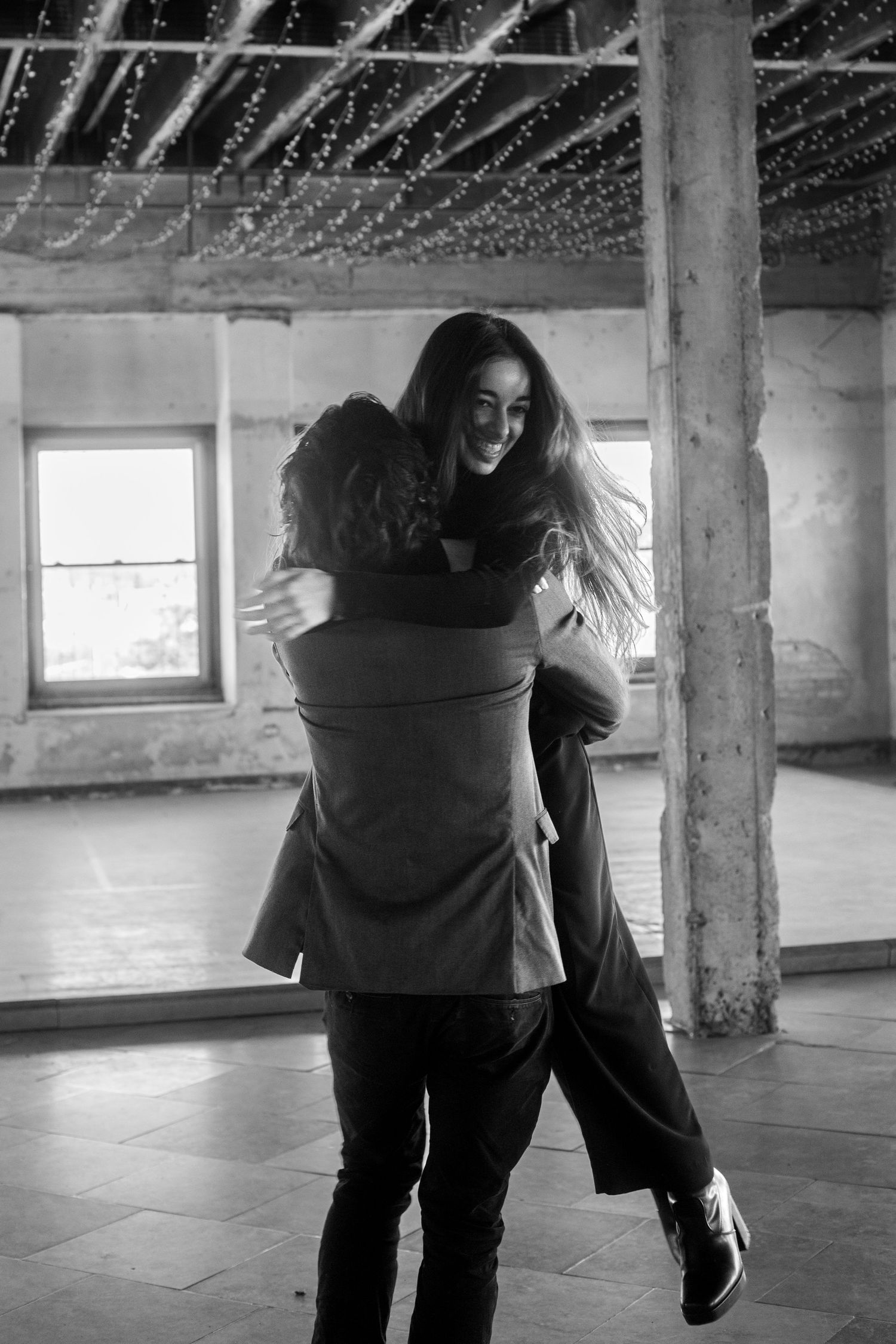 Black and white photo of a man joyfully lifting a laughing woman in an industrial space with string lights.