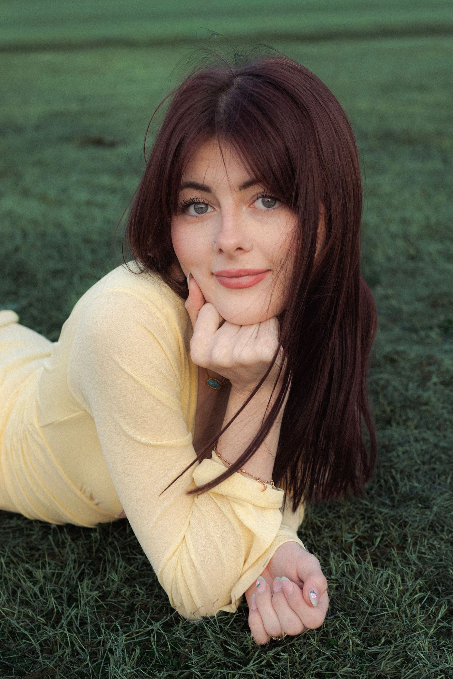 Young woman with auburn hair in yellow dress lying on green grass with hand under chin.