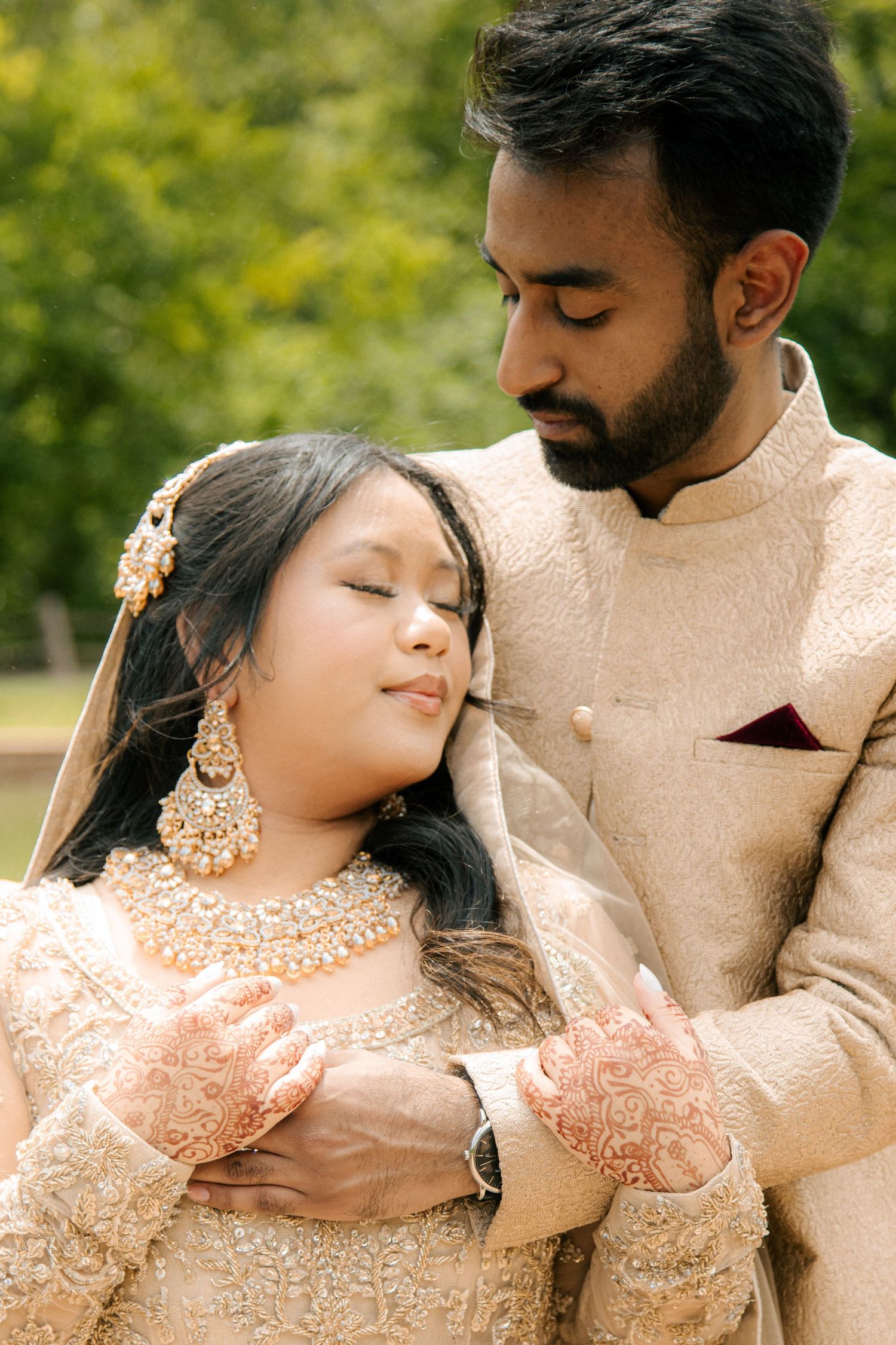 Indian groom in gold sherwani embracing bride in ornate bridal lehenga with henna hands in outdoor setting.